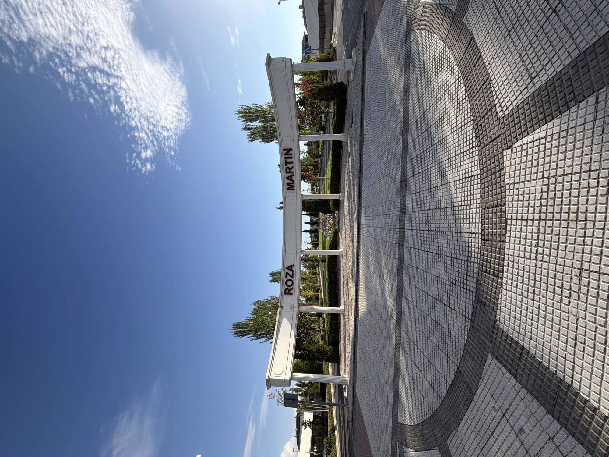 The image depicts an outdoor scene with a clear blue sky and scattered white clouds. In the foreground, a paved area with a mosaic pattern of light and dark square tiles forms a walkway. Behind this, an ornate white archway stands, supported by fluted columns. The words "ROZA MARTIN" are emblazoned in dark red letters on the arch's horizontal beam. Beyond the archway, a green lawn is visible, dotted with trees and shrubs. In the background, buildings with white walls and dark roofs are present, along with a road and a pedestrian crossing sign. The overall impression is of an entrance to a well-maintained public space or a residential area in Majadahonda, Spain, likely during daylight hours given the bright lighting and shadows. No people are visible in the frame.