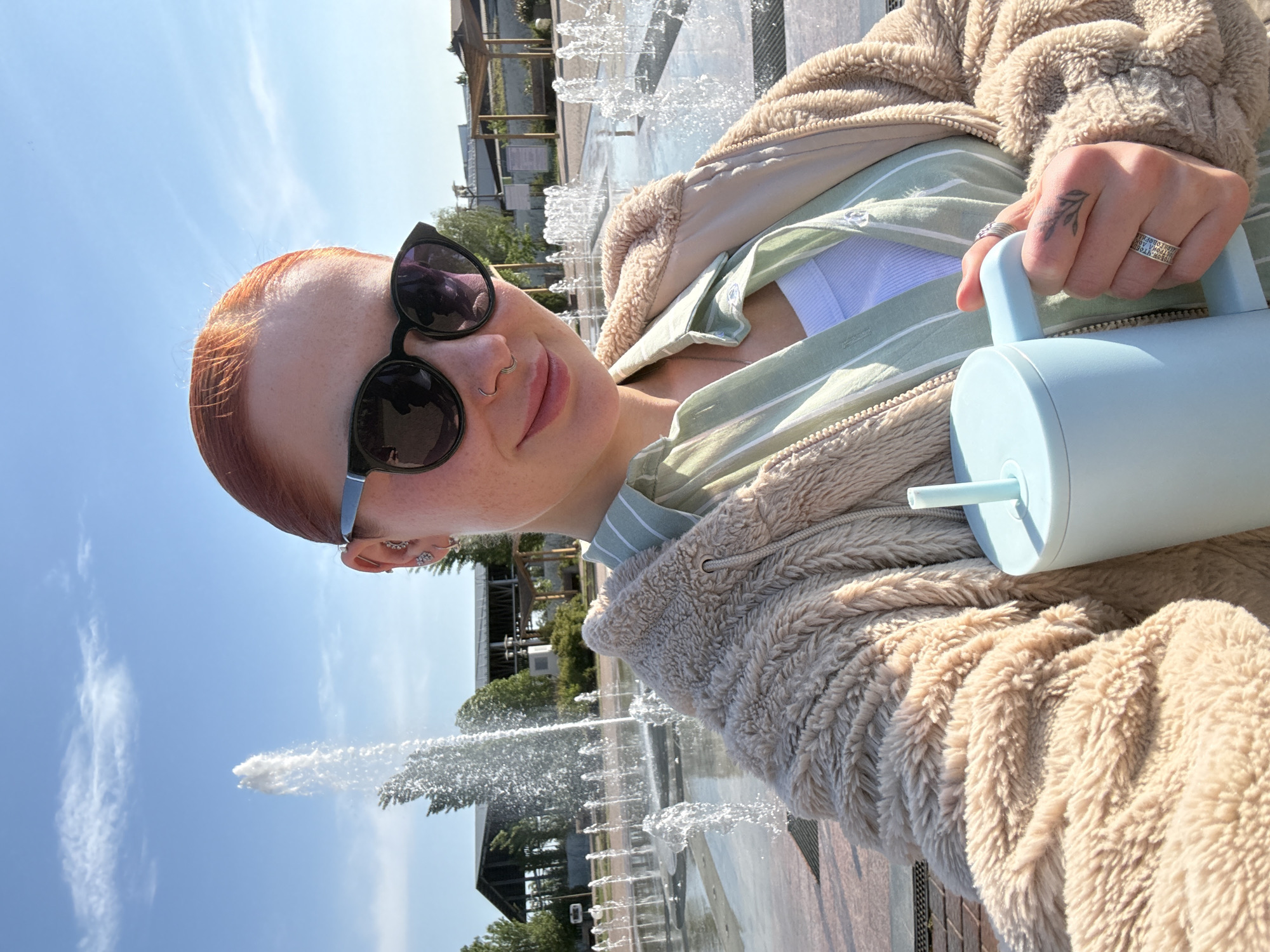A woman with red hair and sunglasses is holding a blue water bottle and posing for a selfie in front of a fountain. The woman is wearing a green striped shirt, a white tank top, and a tan fluffy jacket. The fountain is in a paved area with trees and buildings in the background. The sky is blue. The woman has a tattoo on her left hand. The location appears to be a public space in the city of Rostov-on-Don, Russia, based on architectural styles and common urban features.