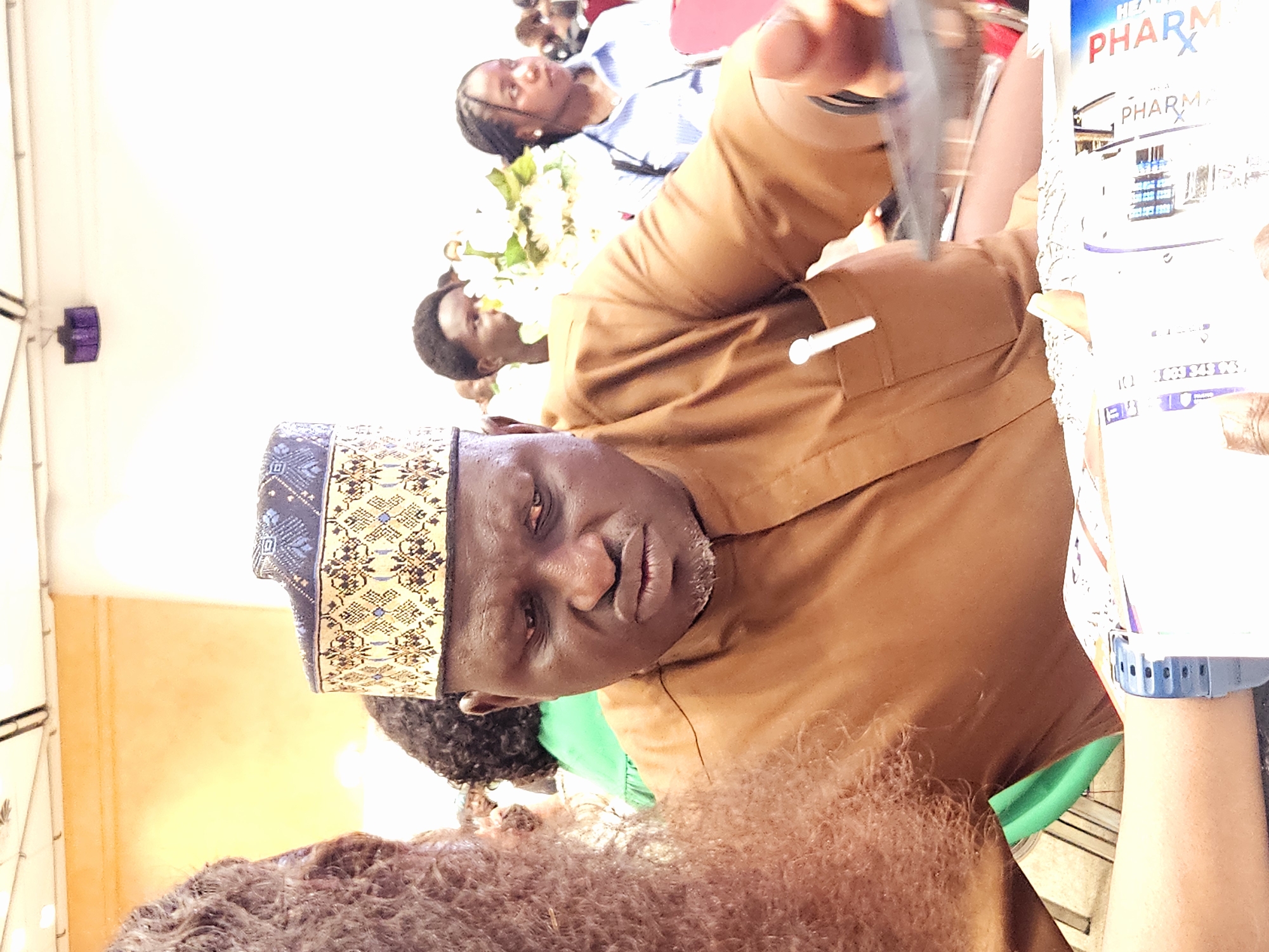 The image shows a close-up of a Black man wearing a patterned kufi and a brown outfit, seated at a table in what appears to be an indoor event. His gaze is directed downwards, and his mouth is slightly ajar. He is holding a transparent object, possibly a phone or tablet, in his right hand. A white pen is clipped to his garment. In the foreground, a portion of a person's arm with a blue digital watch is visible. Other individuals and floral decorations are discernible in the background, suggesting a gathering or conference. The lighting is bright, and the environment seems to be well-lit, possibly a conference hall or banquet room. The image has a slight tilt, with the top of the frame appearing to the left. The overall scene conveys a moment of focus or observation during an event.