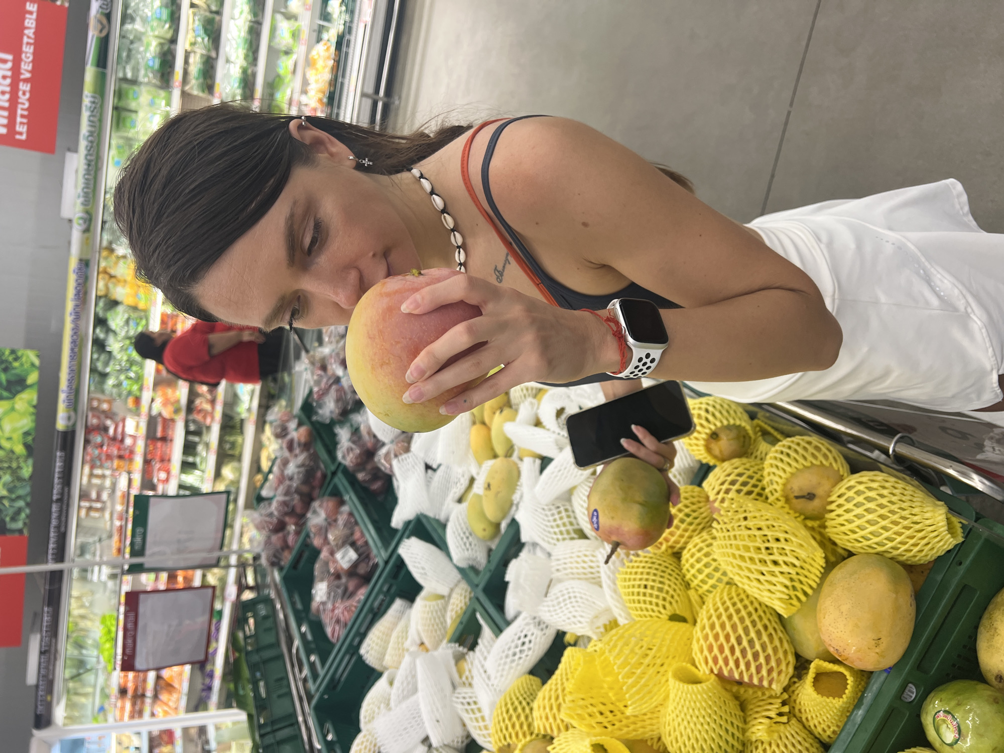 A woman is in a supermarket, likely in Baan Bo Phut, Thailand, considering the Thai writing on signs in the background. She's holding a mango up to her nose, seemingly smelling it. The woman has dark hair, a seashell necklace, and a red string bracelet. She is wearing a smartwatch on her wrist and holding a black smartphone. She is wearing a black and blue tank top with red straps and white shorts. There is a shopping cart to her right that contains a variety of mangoes, some covered in yellow mesh and some covered in white mesh. There are also some dark plums to her left. Supermarket shelves with various vegetables and fruits are visible in the background.
