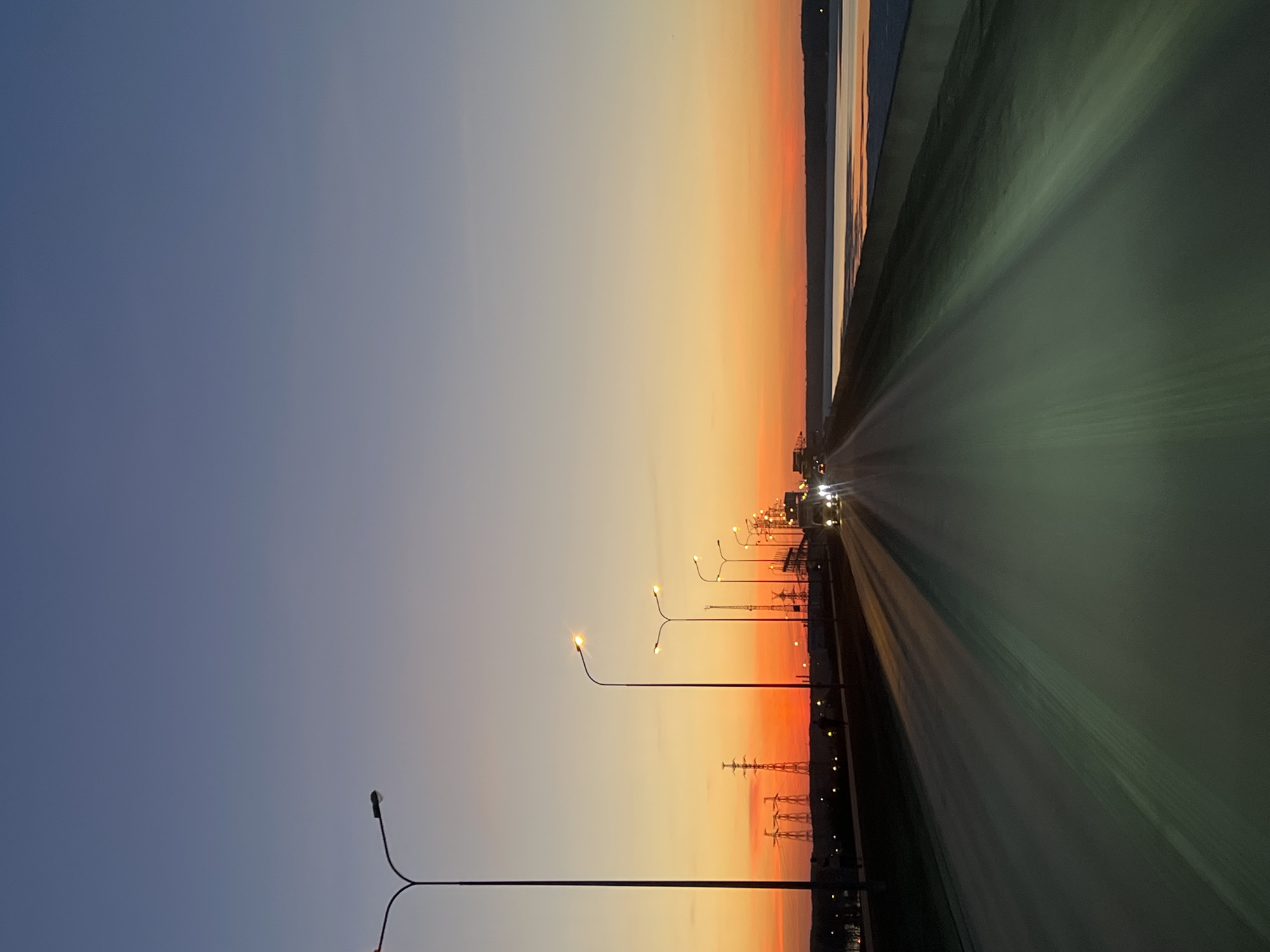 The image displays a wide, expansive view of a road or bridge at either sunrise or sunset, captured in a landscape orientation. The sky occupies the upper portion of the frame, showcasing a dramatic color gradient. It transitions from a deep, muted blue-grey in the upper left, blending through lighter blues and subtle purples in the middle, to vibrant oranges and fiery reds closer to the horizon line on the right.
A clear horizon stretches across the mid-right of the image, separating the colorful sky from the distant landscape. Along this horizon, a dark, undulating silhouette suggests a distant landmass or mountains. To the right of the road, a body of water is visible, reflecting the bright orange and red hues of the sky in horizontal streaks of light.
The lower and right-hand side of the image is dominated by a broad, multi-lane road or bridge that appears to stretch diagonally into the distance. The surface of the road has a slightly greenish-grey tone, possibly due to artificial lighting or ambient reflections. Long, parallel streaks of light on the road surface suggest either a long exposure photograph or the perspective from a moving vehicle, conveying a sense of speed or motion.
Along the right edge of the road, a series of streetlights are illuminated, casting warm, bright yellow-orange glows. Some of these streetlights feature distinct double arms. Further down the road, faint but noticeable headlights from distant vehicles are visible, indicating traffic moving along the path. Below the streetlights and silhouetted against the brightest part of the sky, several electricity pylons or power line towers are present, adding an element of industrial infrastructure to the scene.
The overall impression is one of tranquility blended with the activity of transportation, set against a strikingly colorful natural backdrop. No discernible text is visible in the image.