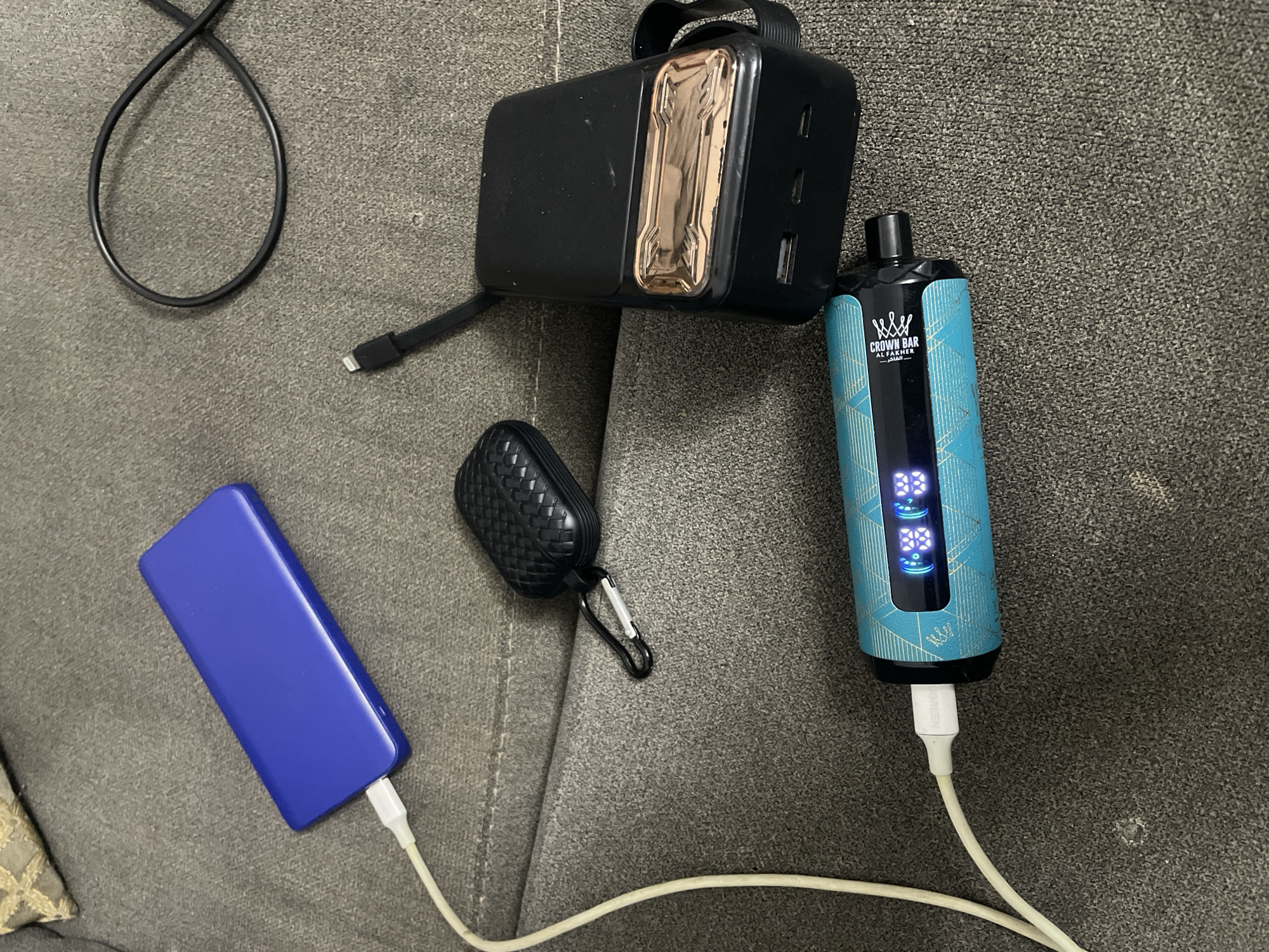 The image displays a close-up, overhead view of various electronic devices resting on a textured gray surface, likely upholstery. In the upper left corner, a bright blue rectangular power bank is connected to a white charging cable. To its right, a black coiled cable lies loose. Below the blue power bank and slightly to the left, a black protective case, possibly for earphones, is visible with a silver carabiner attached. In the center right of the image, a black rectangular power bank with a rose gold accent band is positioned. In the foreground, a teal cylindrical device with a patterned design and a digital display is connected to a white charging cable. The digital display on the teal device shows illuminated icons and numbers, suggesting it is powered on or charging. The scene is devoid of any indication of a specific location in Abuja, Nigeria, or any other geographical context. The overall impression is one of organized clutter of personal electronics.