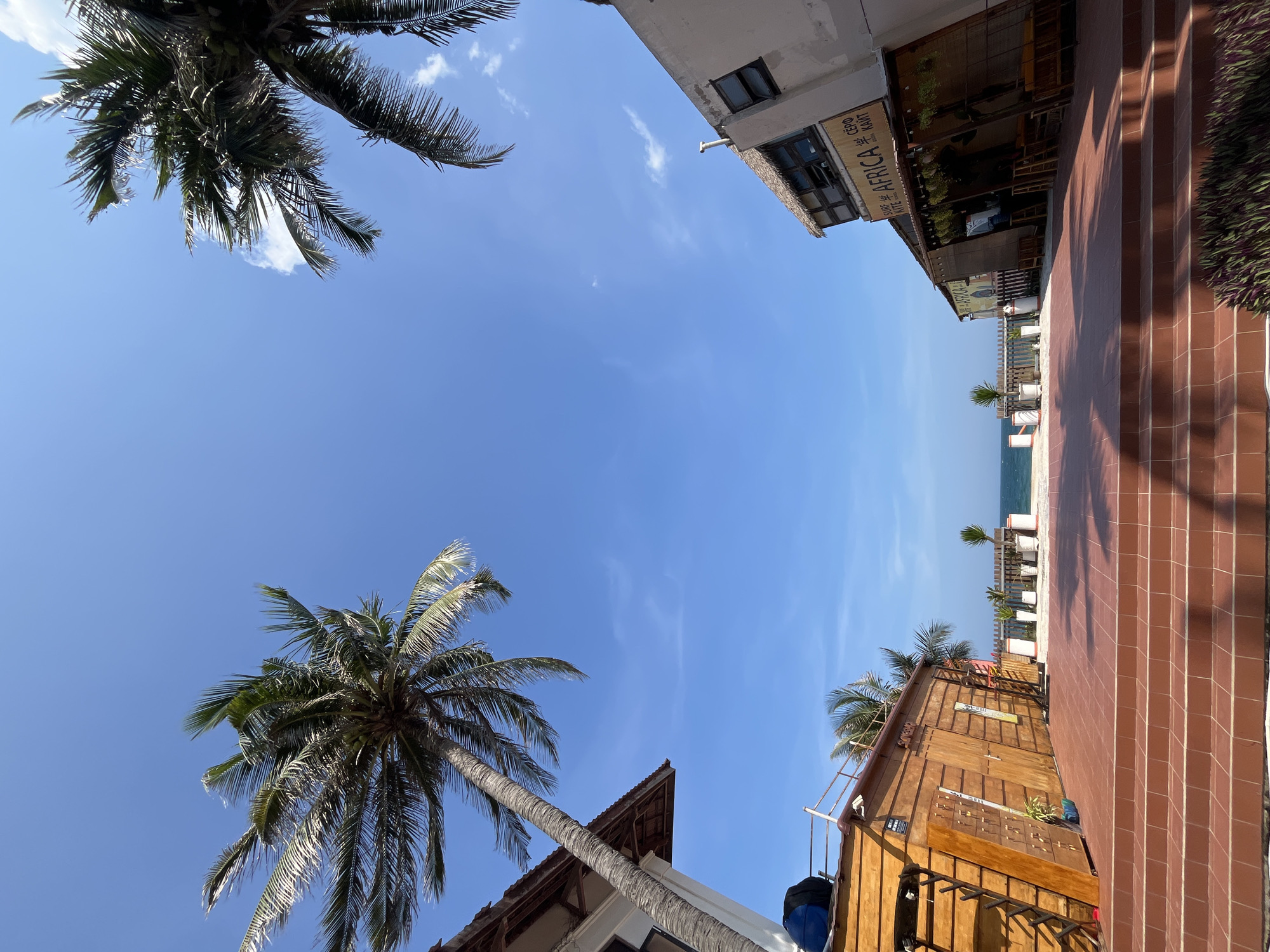 The image depicts an outdoor scene in Phu Thuy Ward, Vietnam, likely a beachside establishment. Two buildings, one on the left with a white facade and a wooden lower section, and another on the right with a thatched roof and a sign reading "SURF KITE AFRICA," flank a tiled patio. Both buildings appear to be open-air establishments with seating visible. Palm trees frame the scene, with their fronds extending into a clear blue sky. In the background, the ocean is visible, separated from the patio by a wooden fence and several potted plants. The overall impression is that of a tropical, relaxed environment, possibly a cafe or surf shop near the coast.