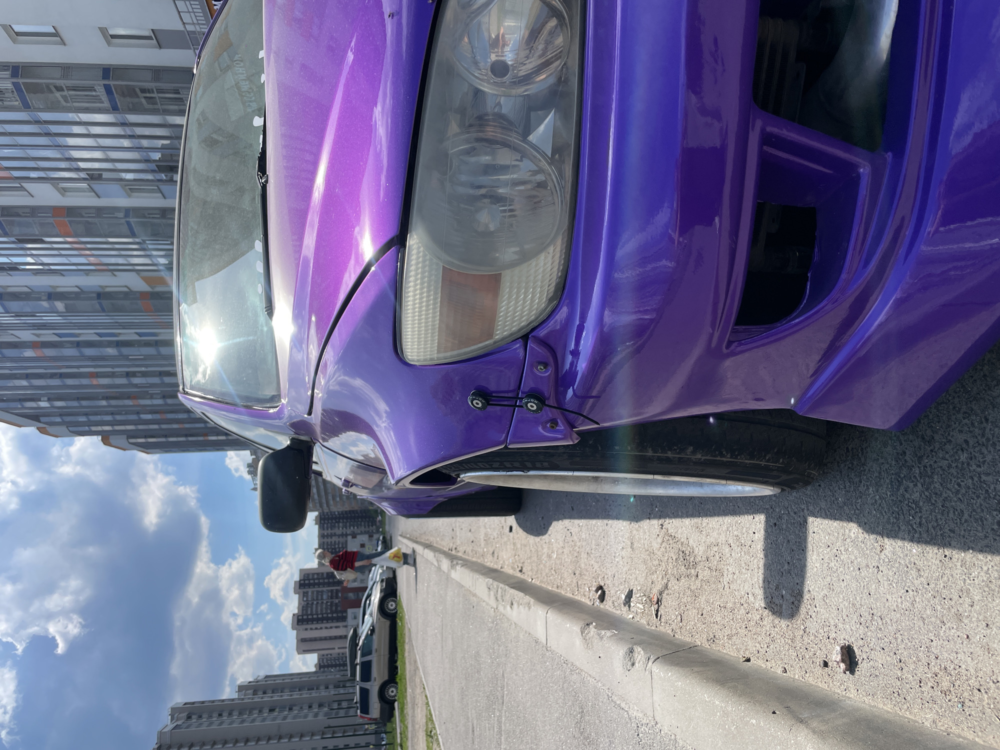 A low-angle, eye-level photograph captures a highly modified purple Lexus IS (XE10 chassis) parked alongside a concrete curb on an asphalt street in a residential area of Unknown, Russia. The vehicle, painted a vibrant metallic purple, features widebody fender flares attached with visible black quick-release fasteners and bungee-like straps, accommodating deeply dished wheels with stretched tires exhibiting significant negative camber. The front bumper is an aftermarket design with prominent lower air intakes. The headlights appear somewhat hazed. The windshield displays dirt and a visible reflection of the sky, with white Russian text "Я СЕМЕЙНОК" (I AM FAMILY MAN) present on its surface.
The sunlight is bright, creating distinct shadows, suggesting midday conditions. The asphalt road surface shows minor wear and scattered small debris near the curb, which is chipped and worn. In the mid-ground, a person, likely female with light-colored hair, wearing a red and white striped long-sleeved shirt, blue jeans, and a backpack, walks away from the camera, carrying a yellow plastic bag in her right hand. Behind her, a beige Volvo V70 station wagon with a roof box and another silver sedan are parked. The background features multiple multi-story residential buildings under a blue sky with scattered white clouds. The buildings vary in style, with some appearing older and others presenting modern reflective glass facades.