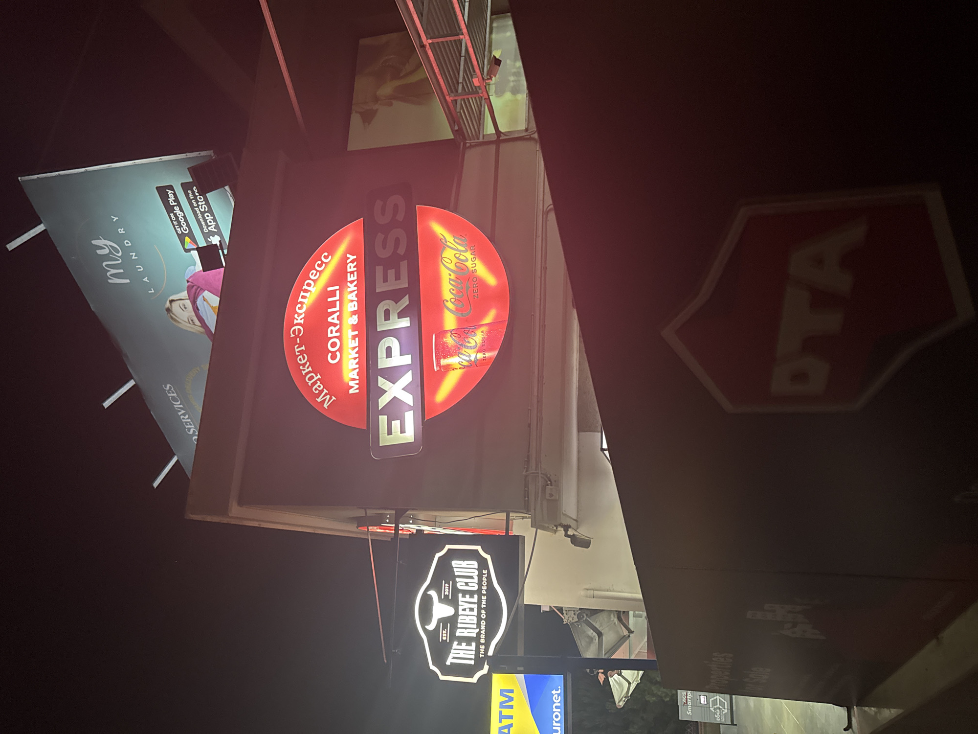 In the city of Agios Tychona Community, Cyprus, at night, a vibrant neon sign for "EXPRESS MARKET & BAKERY" illuminates the scene. The sign is circular and red, with yellow accents and a prominent "EXPRESS" logo in the center. Below it, a red Coca-Cola Zero Sugar can is depicted. To the left, another neon sign reads "THE RIBEYE CLUB" with a bull's head logo and "EST. 2017". Adjacent to this is a bright yellow ATM sign for "euronet.". In the background, a large billboard for "my LAUNDRY" is visible, featuring a woman and app store download icons. Further up, a sign for "ACS Smartparcel Services" is partially visible. The overall environment suggests a commercial area, possibly a street or plaza, with multiple businesses advertising their services through illuminated signage.