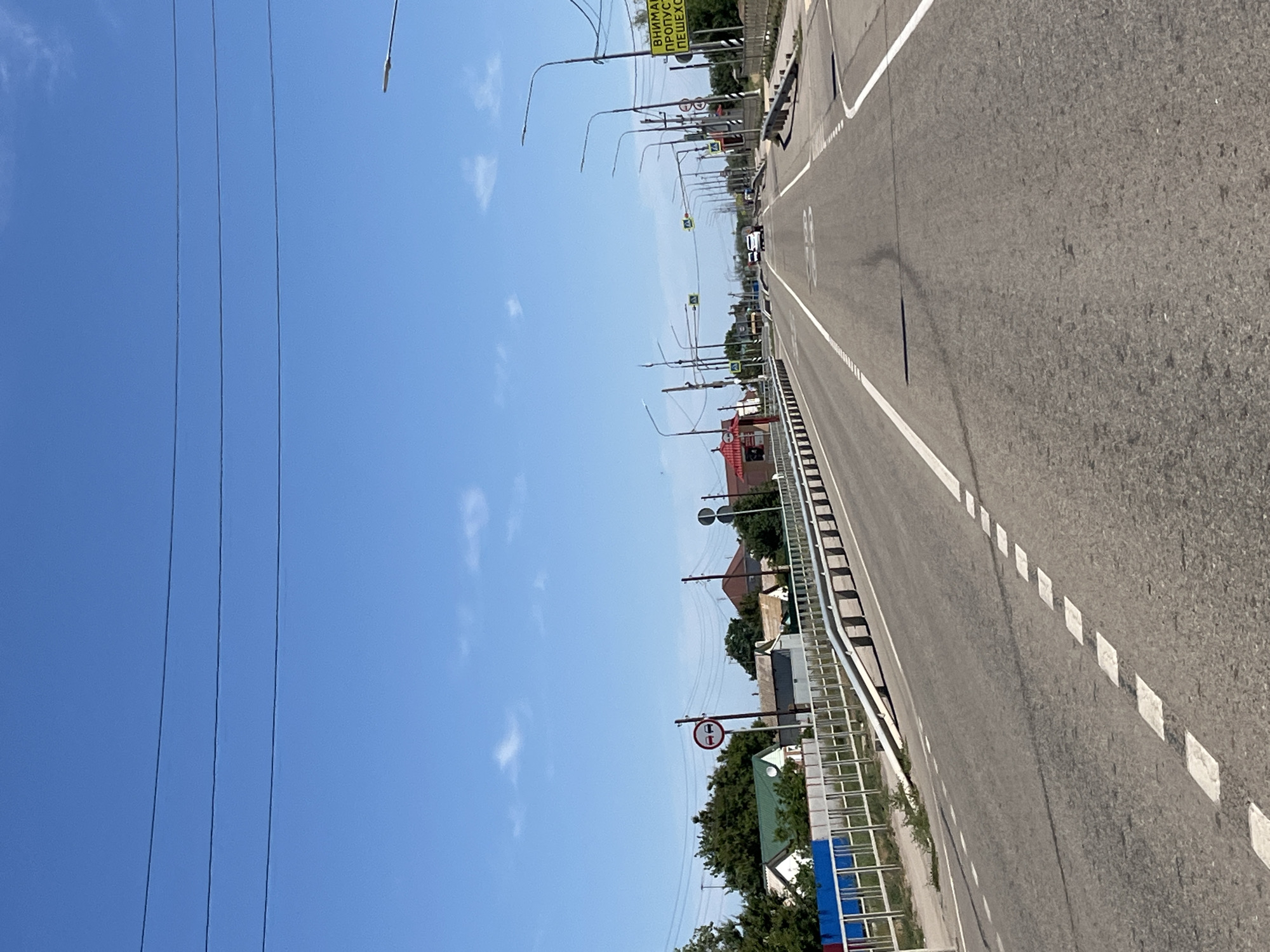 The image shows a straight asphalt road in Bakhtemir, Russia, leading towards a bright horizon under a clear blue sky with scattered white clouds. The road is marked with white dashed lines and a solid white line on the right edge. On the left side of the road, a metal railing runs parallel to the street, separating it from a sidewalk and a row of houses with red and green roofs. Several trees are visible behind the houses, and a blue fence is present in the foreground. On the right side of the road, a series of streetlights are erected at regular intervals, and several traffic signs are visible, including a yellow sign with black lettering and a circular red sign with two cars depicted inside. A few vehicles are present on the road, including a white car in the distance. The scene appears to be a typical suburban or rural road in Russia.