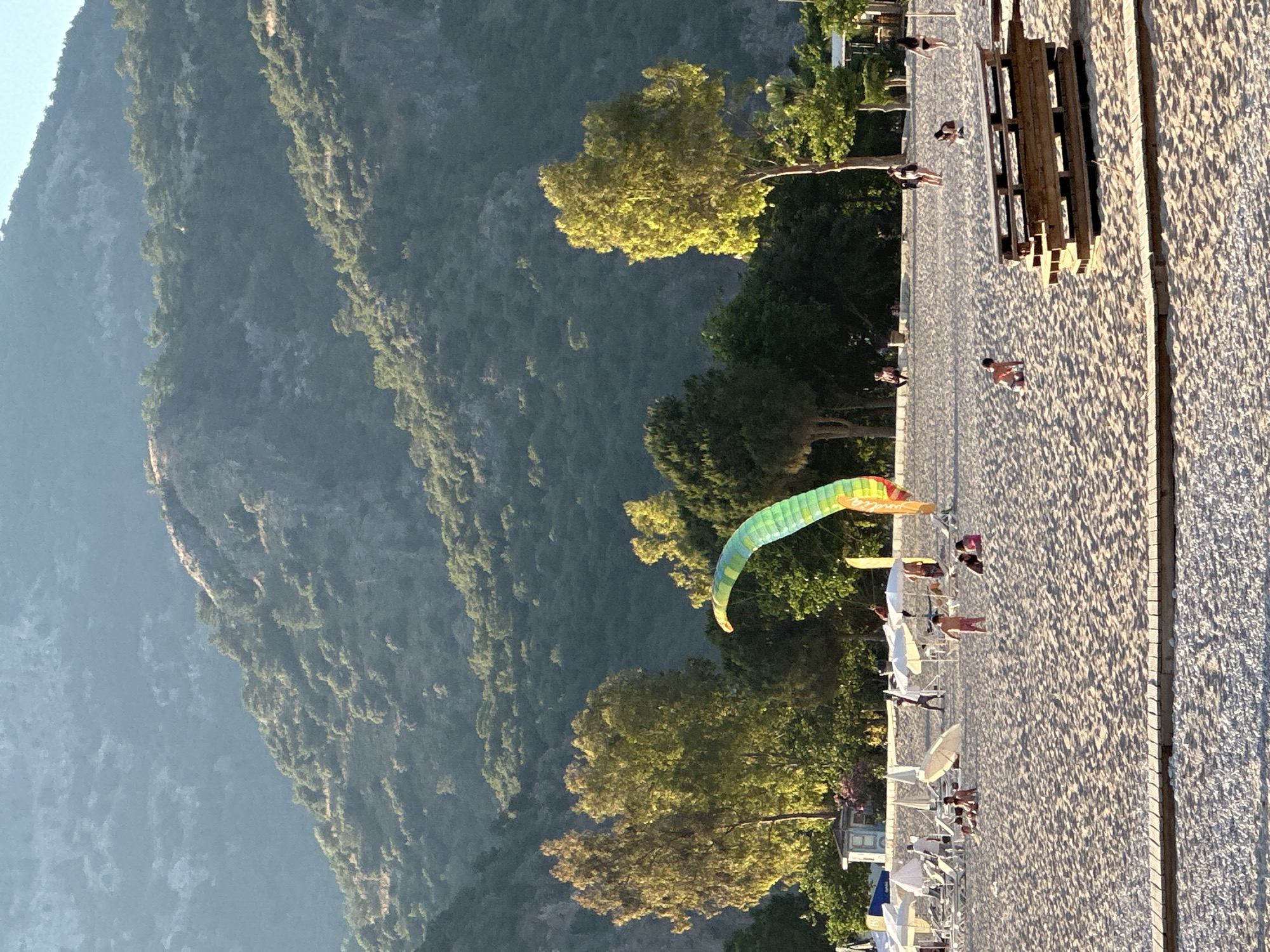The image, when viewed in its correct landscape orientation, captures a vibrant coastal scene in what appears to be Fethiye, Turkey, known for its paragliding. A towering, majestic mountain covered in dense green foliage dominates the left side of the frame, its rugged slopes extending high into the background. The mountain's contours are softened by a slight atmospheric haze, hinting at a clear and expansive sky.
To the right, a wide expanse of light-colored sandy beach stretches across the foreground. Several people are visible on the beach, engaging in leisurely activities. Some individuals are strolling along the shoreline, while others are relaxing on sun loungers or under white beach umbrellas. A prominent wooden structure, possibly a pier, platform, or seating area, is situated on the sand, adding a man-made element to the natural setting.
Along the boundary between the beach and the mountain, a line of verdant trees stands tall, their canopies varying in shades of green, with some brightly illuminated by the sun. Above these trees and over the beach, a colorful paraglider, striped with shades of green, yellow, and orange, gracefully floats in the sky, suggesting an active paragliding site.
The lighting in the scene, characterized by warm, golden hues on the sand and some of the trees, suggests either late afternoon or early morning, creating a peaceful and inviting atmosphere. The weather appears clear and sunny, ideal for both beach activities and paragliding. The presence of the mountain, beach, and paragliding activity strongly points to a location like Oludeniz, near Fethiye. No visible text is present in the image.