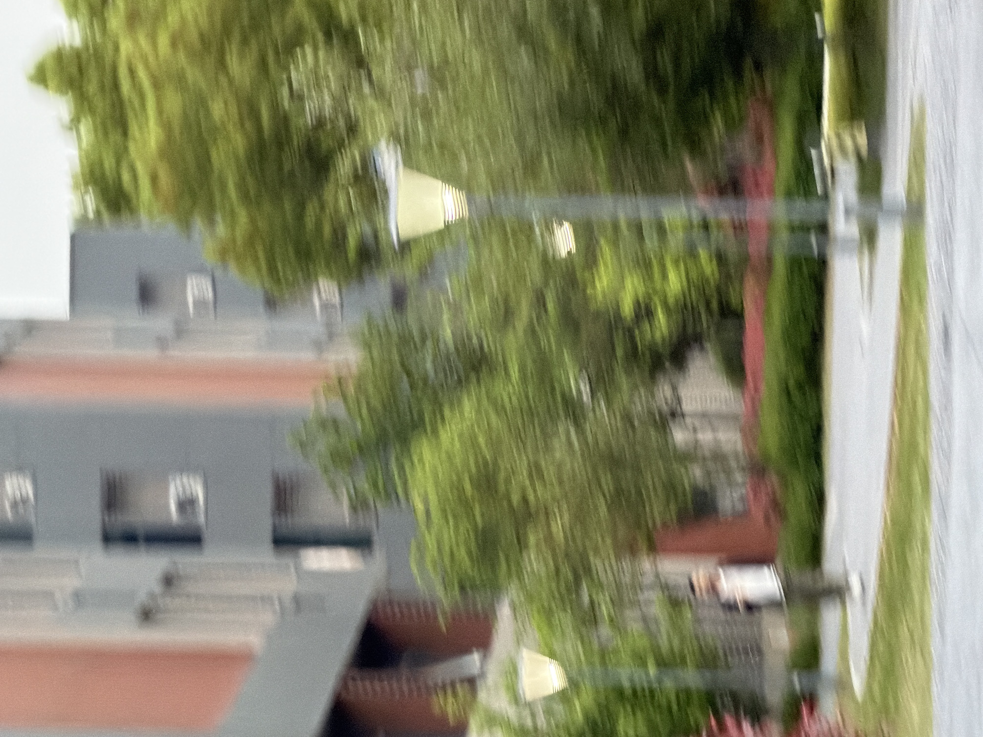 The image shows a blurry, outdoor scene. A multi-story building with a grey facade and horizontal bands of reddish-brown trim is visible. The building has multiple windows. In the foreground, there are trees and greenery. A street lamp stands along a path. The scene appears to be located in an urban or suburban environment. The image does not provide enough detail to pinpoint the exact location in Jiangning District, China.