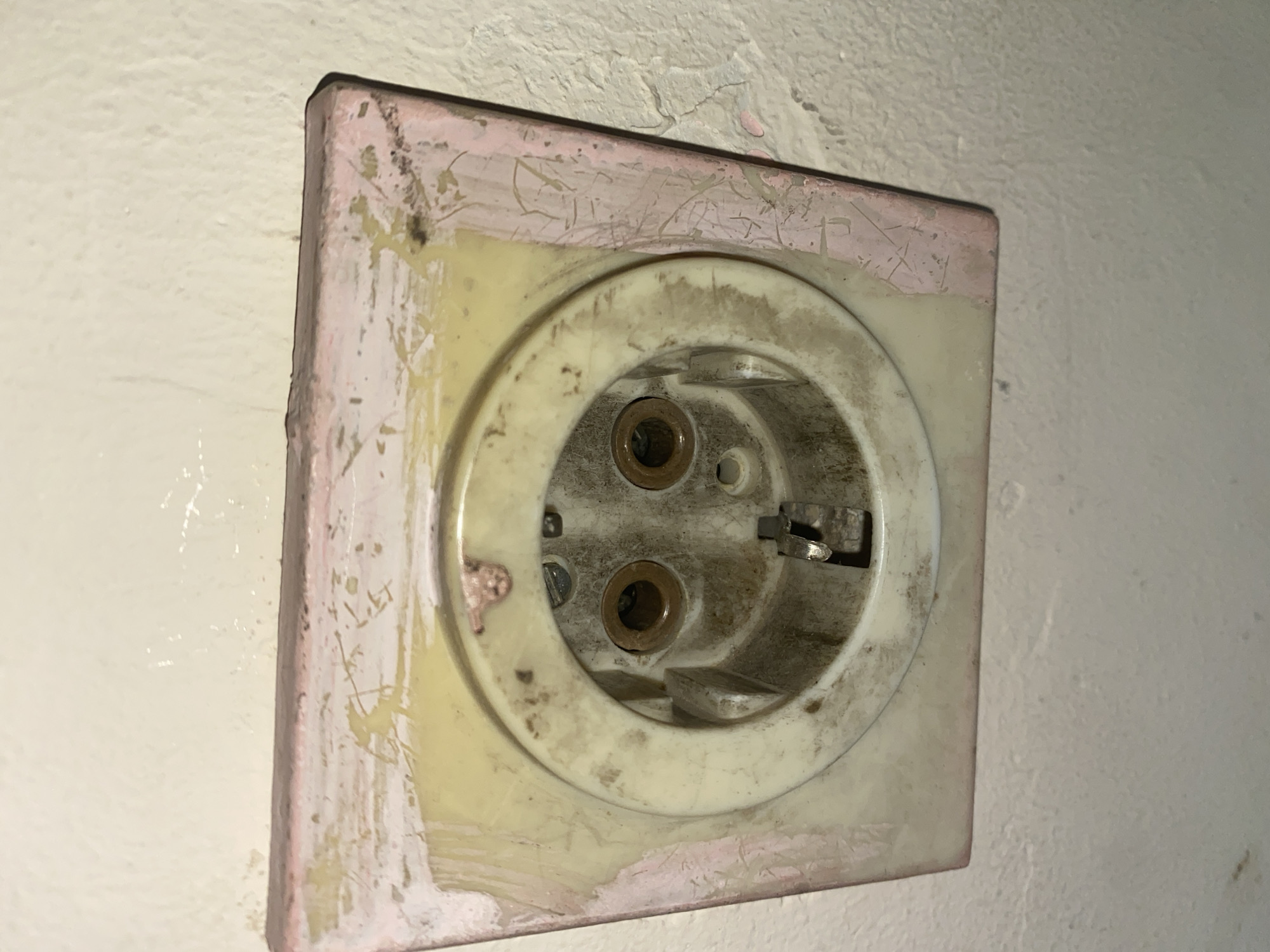 An aged, European-style power outlet is mounted on a textured wall, likely within a building in Sofia, Bulgaria. The outlet, likely a Schuko type, is recessed within a square, potentially painted, plastic or resin housing. The housing exhibits a faded pinkish-brown outer edge and a yellowish inner area around the circular outlet itself. The outlet shows wear and discoloration, with dark deposits or staining visible within the recessed plug receptacles. The wall is painted in a light color.