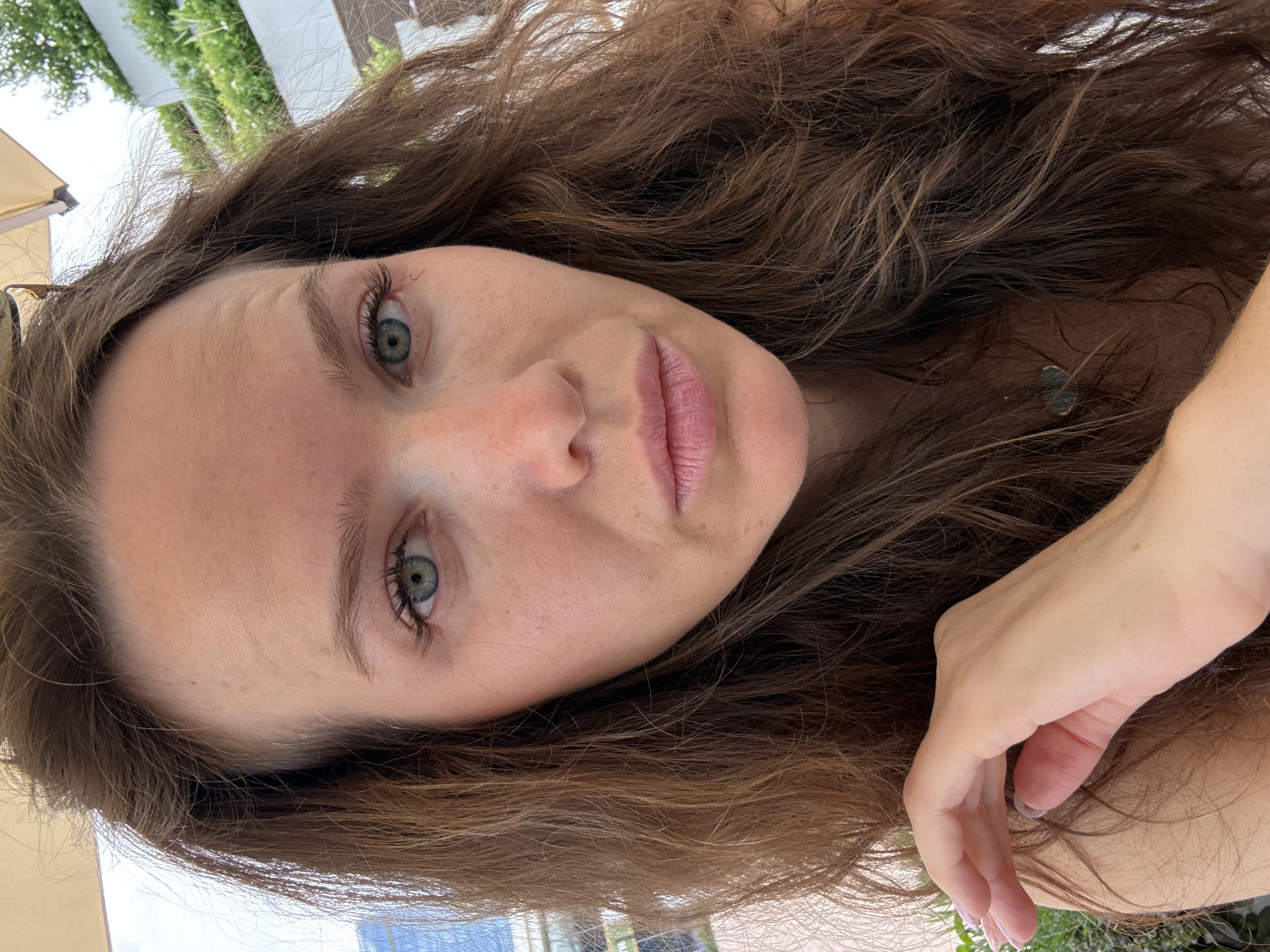 A close-up, tilted portrait captures a woman with long, wavy dark brown hair framing her face. Her light-colored, possibly blue-green, eyes are directed towards the camera with a neutral to serene expression. She has fair skin, a visible smattering of freckles, and natural-looking eyelashes and eyebrows. Her lips are slightly parted, showing a natural pink hue.
Part of a brown or amber-colored sunglasses frame is visible near her left temple. On the right side of the frame, a small, dark, possibly teal-colored earring or part of a necklace is nestled within her hair. A portion of her hand, featuring light-colored nail polish, rests casually on the far right.
The setting is outdoors in Moscow, Russia, during the daytime. The lighting is bright but diffused, suggesting an overcast sky or a shaded area. In the background, hints of green foliage from trees or bushes are visible, alongside white vertical structures, possibly a fence or part of a building. A tan or cream-colored fabric, likely an awning or umbrella, is also discernible in the upper left. No text is visible in the image. The overall impression is a casual, intimate portrait taken in an outdoor environment such as a garden or patio.