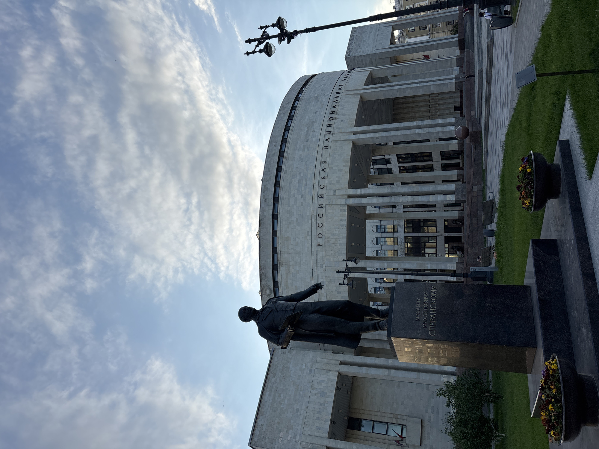 The image, taken with an upward-angled perspective and rotated ninety degrees clockwise, presents an outdoor scene in Saint Petersburg, Russia. In the immediate foreground, a dark bronze statue of a man stands on a robust black granite pedestal. The statue depicts a man in a coat, with his left arm extended and his right hand appearing to hold a book or document. The inscription on the pedestal identifies the subject as "МИХАИЛУ МИХАЙЛОВИЧУ СПЕРАНСКОМУ" (To Mikhail Mikhailovich Speransky).
Behind the statue is a large, impressive building featuring a curved facade made of light-colored stone. The building has prominent vertical columns and numerous windows. Vertically inscribed along its upper curved section is the text "РОССИЙСКАЯ НАЦИОНАЛЬНАЯ БИБЛИОТЕКА" (Russian National Library), identifying the structure.
A tall, dark street lamp with several ornate light fixtures is visible towards the right of the building. The ground level features paved areas, vibrant green grass, and a dark planter filled with colorful flowers near the base of the statue. The sky above is bright, characterized by soft, wispy white clouds. The lighting suggests it is daytime, possibly late afternoon, creating a clear but gently illuminated atmosphere for this significant historical and architectural landmark. No specific activity or event is taking place; the scene is static.