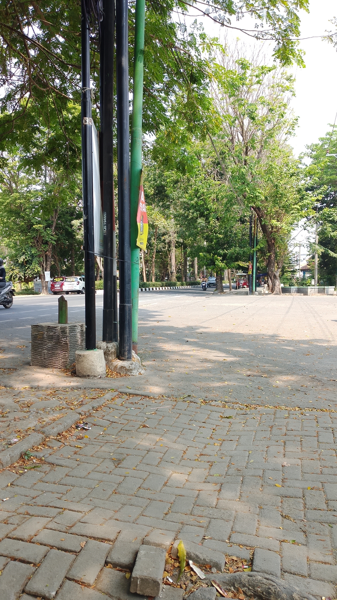 The image captures a street scene in Pakembaran, Indonesia, characterized by a paved walkway in the foreground and a road in the mid-ground. Several dark vertical poles are prominent on the left side of the image, with a green pole situated to their right. Behind the poles, a white car and a red car are visible parked on the roadside, with a motorcycle in motion to the left, and another motorcycle with a rider on the right side of the road. Trees with lush green foliage line the street, creating a shaded environment. The walkway is constructed of gray interlocking pavers, some of which are slightly displaced, with fallen leaves scattered across the surface. In the background, more trees and some buildings are visible, suggesting a well-treed urban or suburban area. The sky is bright and appears to be a clear day.