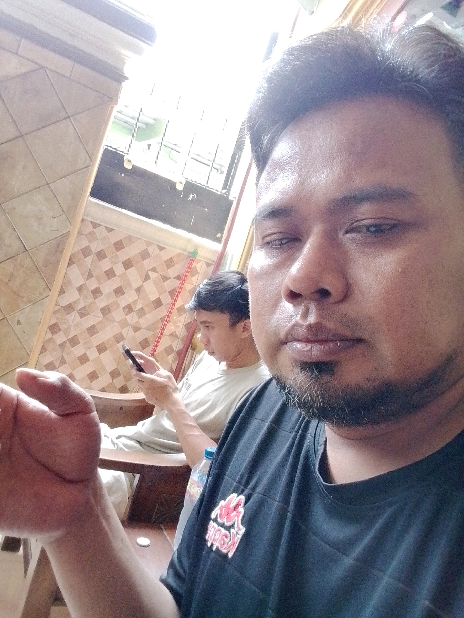 This is a selfie taken by a man in Babelan, Indonesia. The man has dark skin, dark hair, and a goatee. He is wearing a black t-shirt with a red and white logo. He is looking at the camera with his eyes partially closed. In the background, another man is sitting and looking at his phone. The setting appears to be a room with tiled walls and a window with bars. There is a bottle of water on a table in the foreground. The lighting is bright, suggesting it is daytime.
