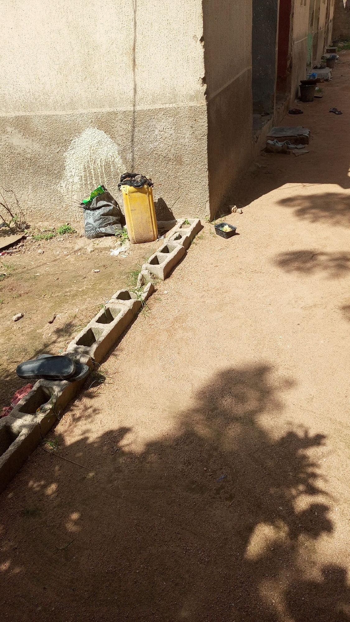 An outdoor scene in Jalingo, Nigeria depicts a dusty, sandy ground with sparse vegetation. In the background, a building with a textured, cream-colored wall stands prominently. A large yellow plastic container is placed near the building, and next to it is a black garbage bag. A wall with a water stain or marking is visible on the building's surface.
In the foreground and middle ground, a series of hollow concrete blocks are laid out in a line on the ground. A pair of black flip-flops rests on top of one of these blocks. Further along the row of blocks, a small black plastic container with some yellow contents is visible. The ground is a mixture of sand and small gravel. Shadows of tree branches and leaves are cast across the sandy ground, indicating it is daytime. Other buildings and doorways are visible in the background, suggesting a residential or commercial area. A few other scattered objects, possibly other containers or debris, are present further down the path.