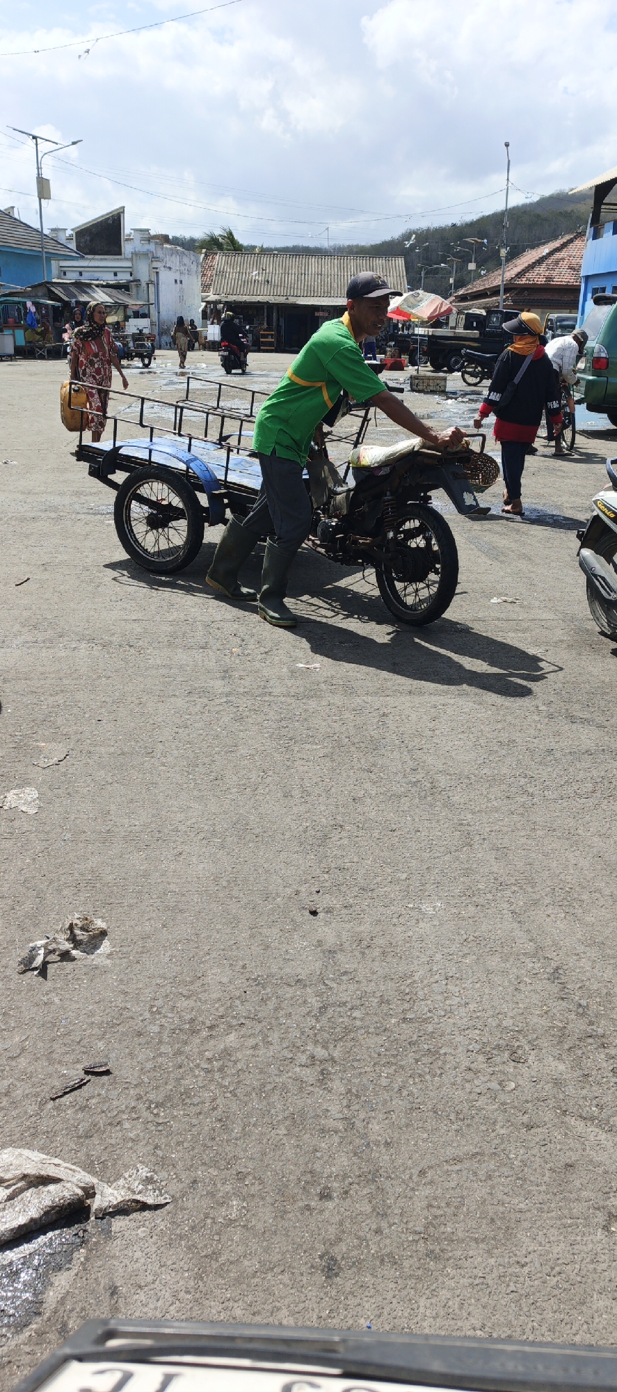 A man in a green shirt and dark pants pushes a motorized cart with a trailer. The cart is attached to a motorcycle. The man is wearing tall green rubber boots. The scene is outdoors, with a paved surface. In the background, there are buildings, other people, and vehicles, including motorcycles and a truck. The sky is partly cloudy. The location is identified as Puger, Indonesia.