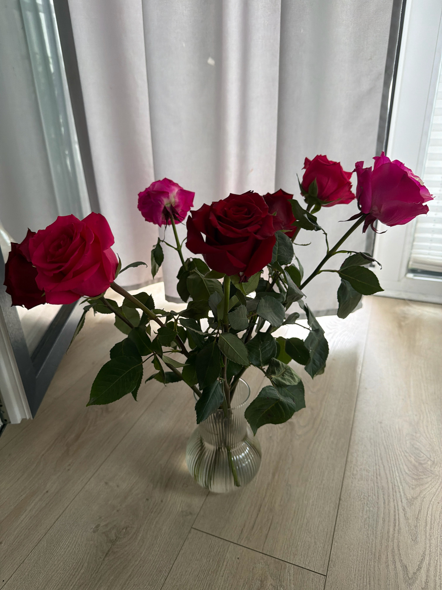 A bouquet of red and pink roses is in a clear glass vase on a light-colored wooden floor. The roses are arranged on thin green stems with dark green leaves. In the background, a white curtain hangs behind the vase, and to the right, a window with white blinds is visible. The scene suggests an indoor setting, possibly a room with natural light. The specific location in Wrocław, Poland, cannot be determined from the provided image.