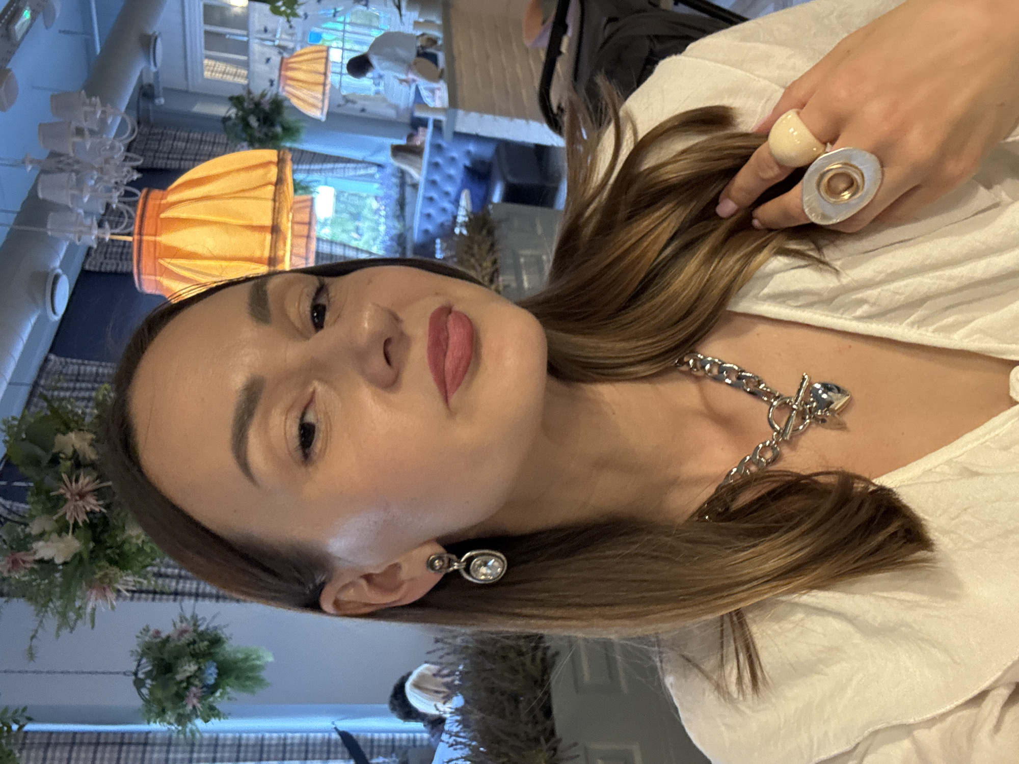 A light-skinned woman with brown hair is the focus of the image, likely taken indoors. She is wearing a white top and noticeable jewelry, including a silver chain necklace with a pendant, matching earrings, and large rings on her fingers.
The background reveals a restaurant interior, with decorative elements like hanging floral arrangements and unique light fixtures. Other people are present in the background, though they are not the central focus.
Specific location in Sochi, Russia, cannot be determined from the image alone.
