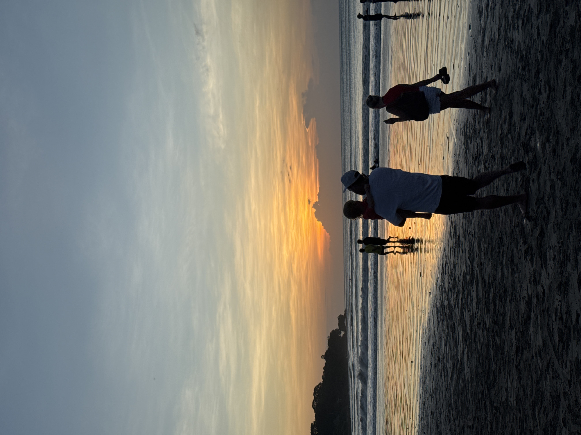 The image shows a sunset scene at a beach in Kuta Selatan, Indonesia. The sky transitions from blue to orange and yellow near the horizon, with clouds streaking across the sky. Silhouettes of people are visible on the beach, including a man holding a child, and other individuals standing or walking near the water's edge. The beach has dark sand and gentle waves reflecting the colors of the sunset.