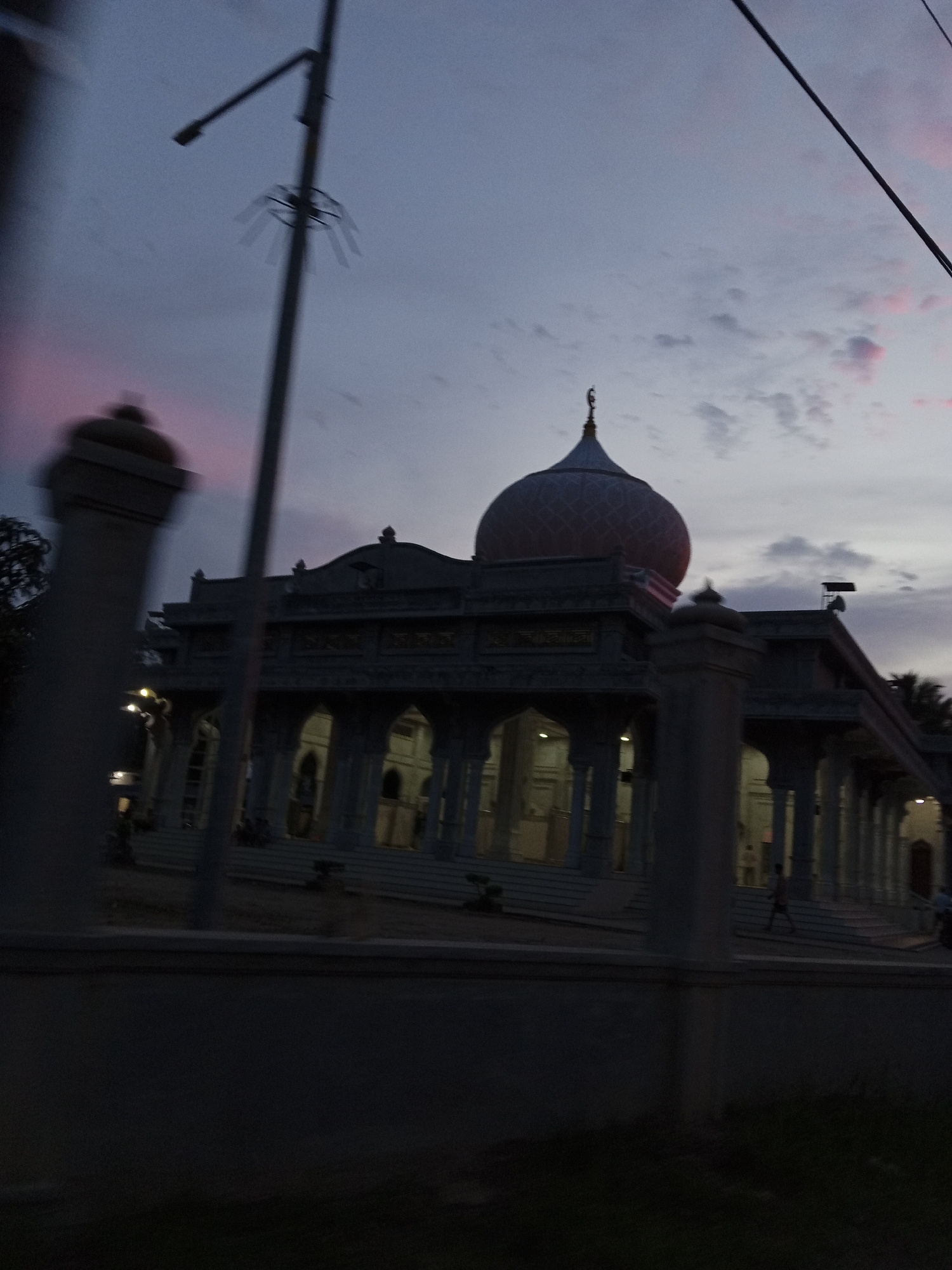 This media file depicts a mosque in Pantebaroglesiblah, Indonesia, at dusk. The sky is a dusky purple with hints of pink from the setting sun, and scattered clouds are visible. The mosque itself is a prominent structure with a large dome topped with a crescent moon finial. The building has a series of arches supported by columns, and lights are illuminated within, suggesting it is evening.
In the foreground, a streetlight with a circular fixture is visible, along with a power line stretching across the top of the frame. The overall scene is somewhat blurry, possibly due to motion blur or a slight out-of-focus capture. A low wall or barrier runs horizontally across the lower portion of the image, separating the foreground from the mosque's grounds.
There are a few indistinct figures of people visible on the steps leading up to the mosque, and one person is walking in the middle ground. The mood of the image is serene and contemplative, fitting for a religious site at the end of the day. No visible text is present in the image.