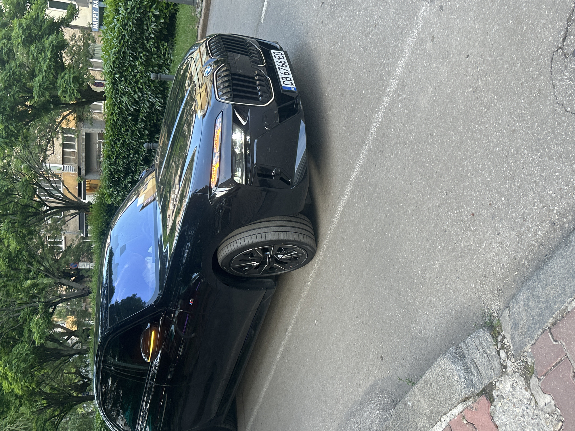 A black BMW 7 Series (G70 generation) is parked or moving slowly on an asphalt road in Sofia, Bulgaria. The vehicle's front right turn signal and the indicator on the right side mirror are illuminated orange. It features a prominent kidney grille, split headlights, and multi-spoke black alloy wheels. A small "M" badge is visible on the front fender. The license plate, "CB 6766 EO," clearly indicates registration in Sofia, Bulgaria. The car's windows are darkly tinted, obscuring interior details.
The road surface is gray asphalt with a faded white dashed line running parallel to the car. A concrete curb separates the road from a sidewalk area. The sidewalk consists of red interlocking pavers near the curb, transitioning to broken concrete and exposed dirt where small weeds are growing.
In the background, a row of mature, leafy green trees lines the street. Behind the trees and a well-maintained green hedge, multiple urban buildings are visible. These include older, multi-story structures with various window designs, some exhibiting a yellowish or light-colored facade. To the far right, a partial sign with the text "HAPPY DA" is discernible. Black bollards are intermittently placed along the edge of the hedge. A round red and white traffic sign is partially obscured by foliage. The scene is illuminated by natural light, suggesting daytime conditions.