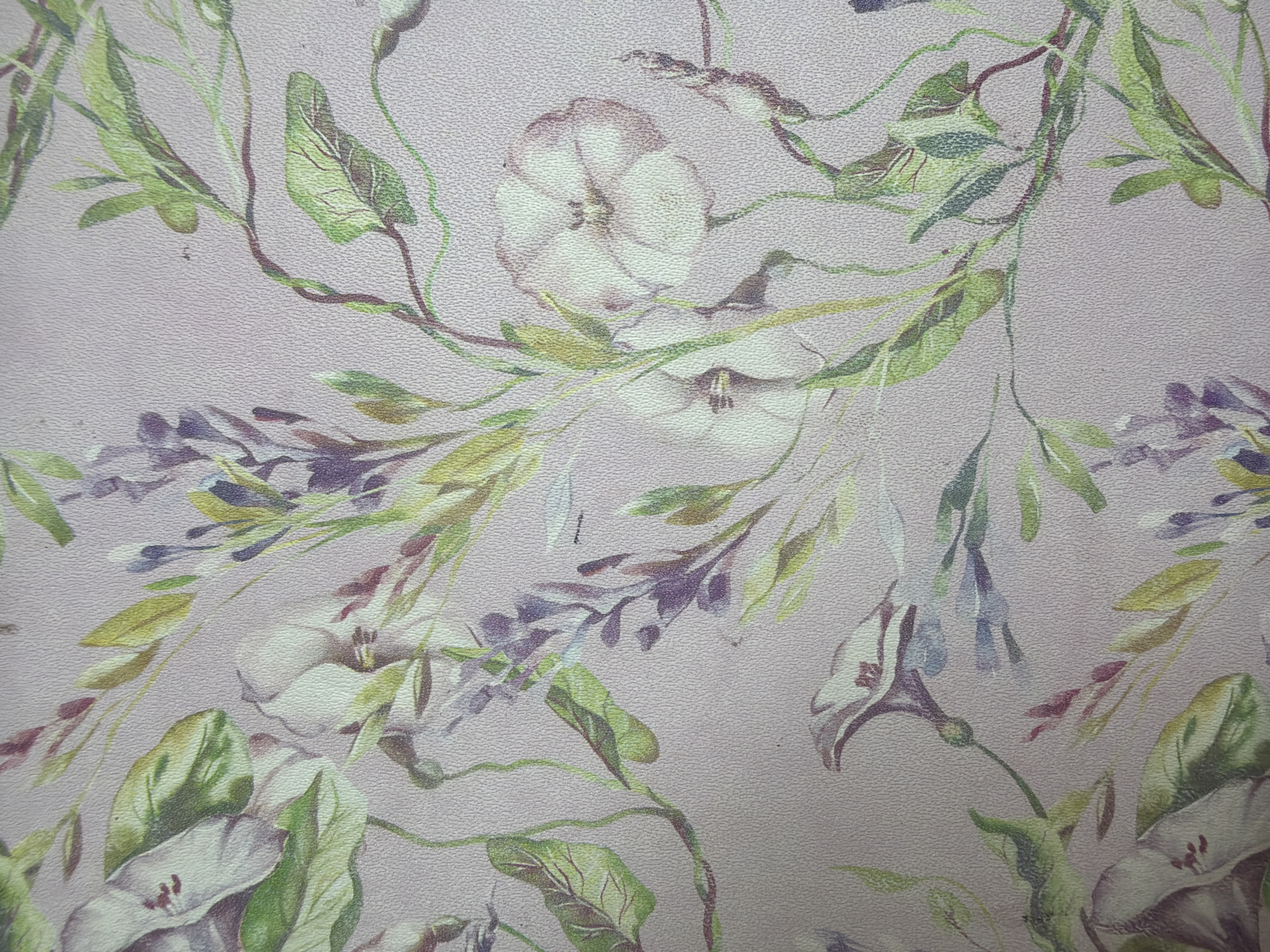 This image is a close-up, flat lay photograph of a floral wallpaper pattern. The pattern features watercolor-style depictions of flowers and foliage in shades of white, pink, purple, and green against a pale pink textured background. There are no people, objects, actions, or interactions visible in this image, nor does it depict any location in Yuzhno-Sakhalinsk, Russia. The image solely presents a decorative surface.