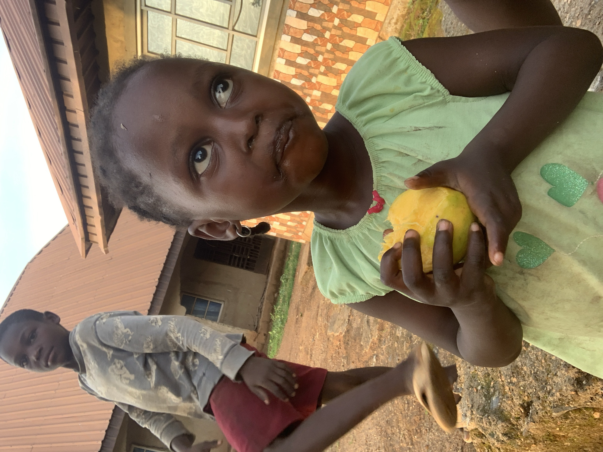 A young child with dark skin is holding a yellow mango in their hands. The child wears a light green dress with heart-shaped patches. The child is looking up and to the side. In the background, another child is standing near a building with a brown roof. The scene is in an outdoor setting. The location is Agban, Nigeria.