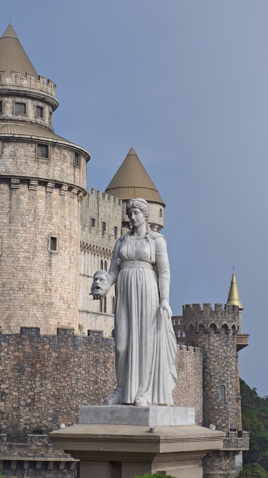 A prominent white marble statue of a woman stands in the foreground, gazing slightly towards the right, against a backdrop of a grand castle complex under a clear, light blue sky. The classical-style statue depicts a female figure clad in a long, flowing gown, with a calm and stoic expression. In her left hand, she holds a detailed, severed male head, characterized by a beard, an open mouth, and wide eyes, possibly representing a theatrical mask of tragedy or a defeated adversary.
Behind the statue, the impressive stone architecture of a castle unfolds. The complex consists of multiple round towers and robust stone walls, adorned with battlements. The towers are capped with distinctive conical roofs; most are a dull bronze or brown, while a smaller spire on the far right shimmers with a bright golden hue. The stonework of the castle walls varies, from lighter, dressed blocks on the larger towers to rougher, darker masonry on the lower walls.
The clear, cloudless sky indicates daytime, likely late morning or early afternoon. A hint of green foliage is visible on the far right, suggesting the castle is situated on a hill or amidst natural surroundings. The scene presents a blend of classical artistry and medieval-inspired architecture, characteristic of themed attractions found in locations like Đà Nẵng, Vietnam. No discernible activity or visible text is present in the image.