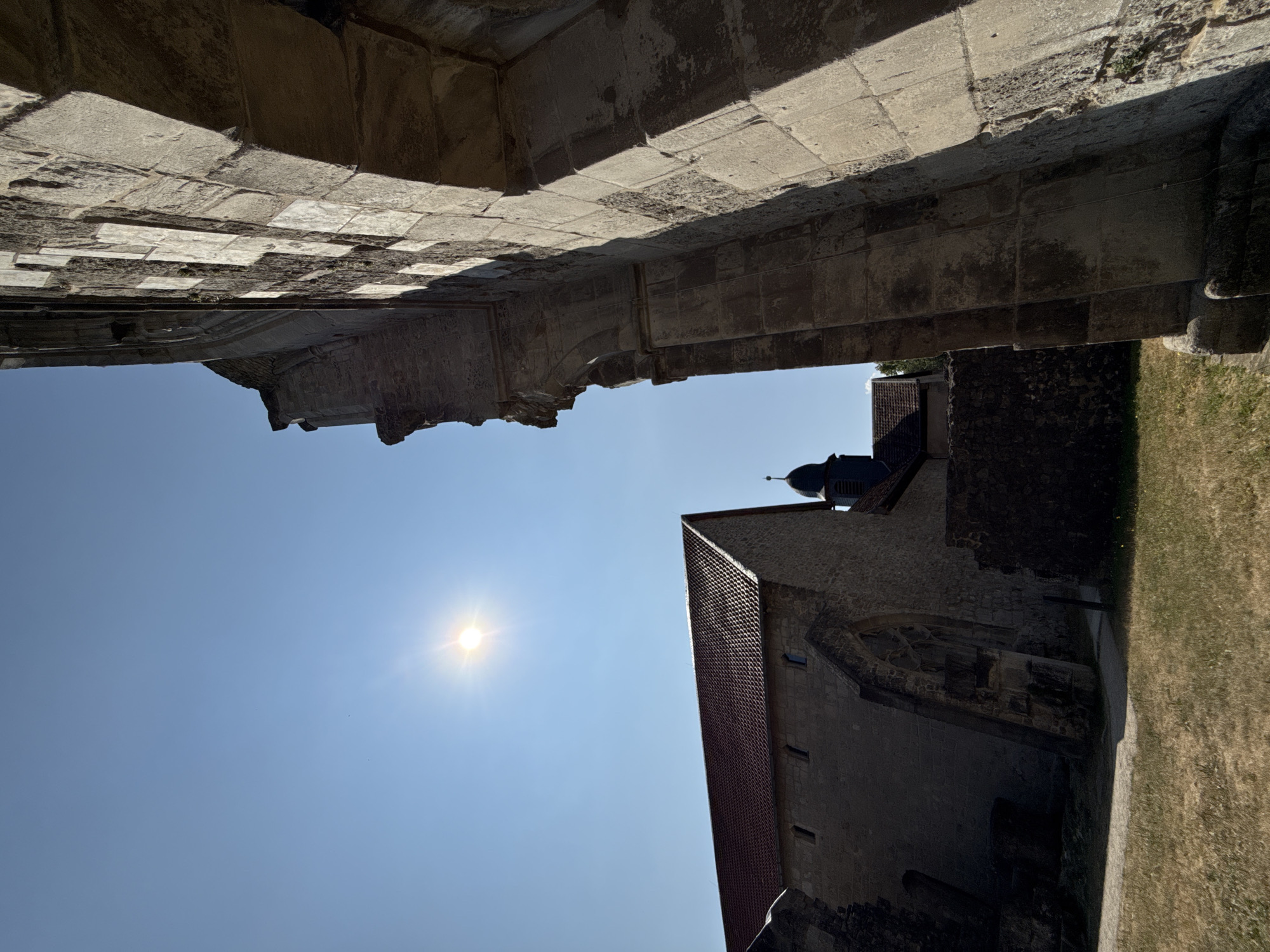 The image shows the ruins of Walkenried Abbey, located in Walkenried, Germany. The sun is visible in the upper left quadrant of the image, shining brightly against a clear blue sky. In the foreground, on the right, is a large stone structure with arched openings and rough-hewn blocks, casting a strong shadow. To the left, a stone building with a red tiled roof is visible, featuring arched windows. A dark spire of a smaller building rises above the roofline behind the main stone structure. The ground is covered with dry grass and some patches of dirt. The scene appears to be a historic site bathed in sunlight.
