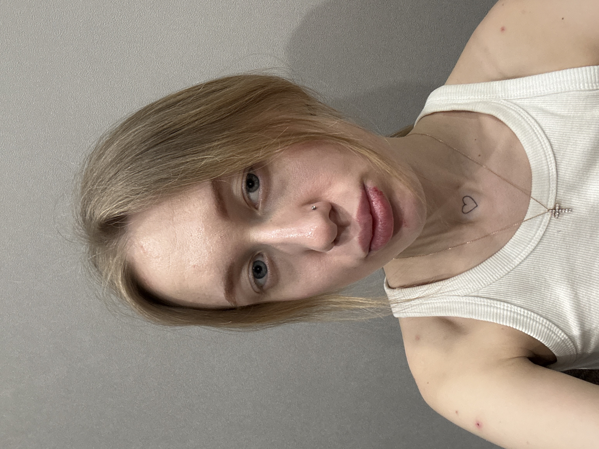 The image depicts a close-up selfie of a young adult female indoors. The exact location in Rostov-on-Don, Russia, cannot be determined from the visual evidence, which shows only a plain, light grey, textured wall as the background.
The subject is a light-skinned female with blonde hair, parted off-center, with some strands falling across her face. She has blue eyes and a small, silver-colored stud piercing on the left side of her nose. Her lips are slightly pursed, and a reddish lesion or sore is present on the right side of her lower lip. Several small, reddish marks or blemishes are visible on her left shoulder and upper arm. A small, black outline of a heart is tattooed on her upper chest, below the left clavicle. She is wearing a white, ribbed tank top and a thin, light-colored chain necklace with a small, cross-shaped pendant adorned with clear stones. She is looking directly at the camera with a neutral expression.