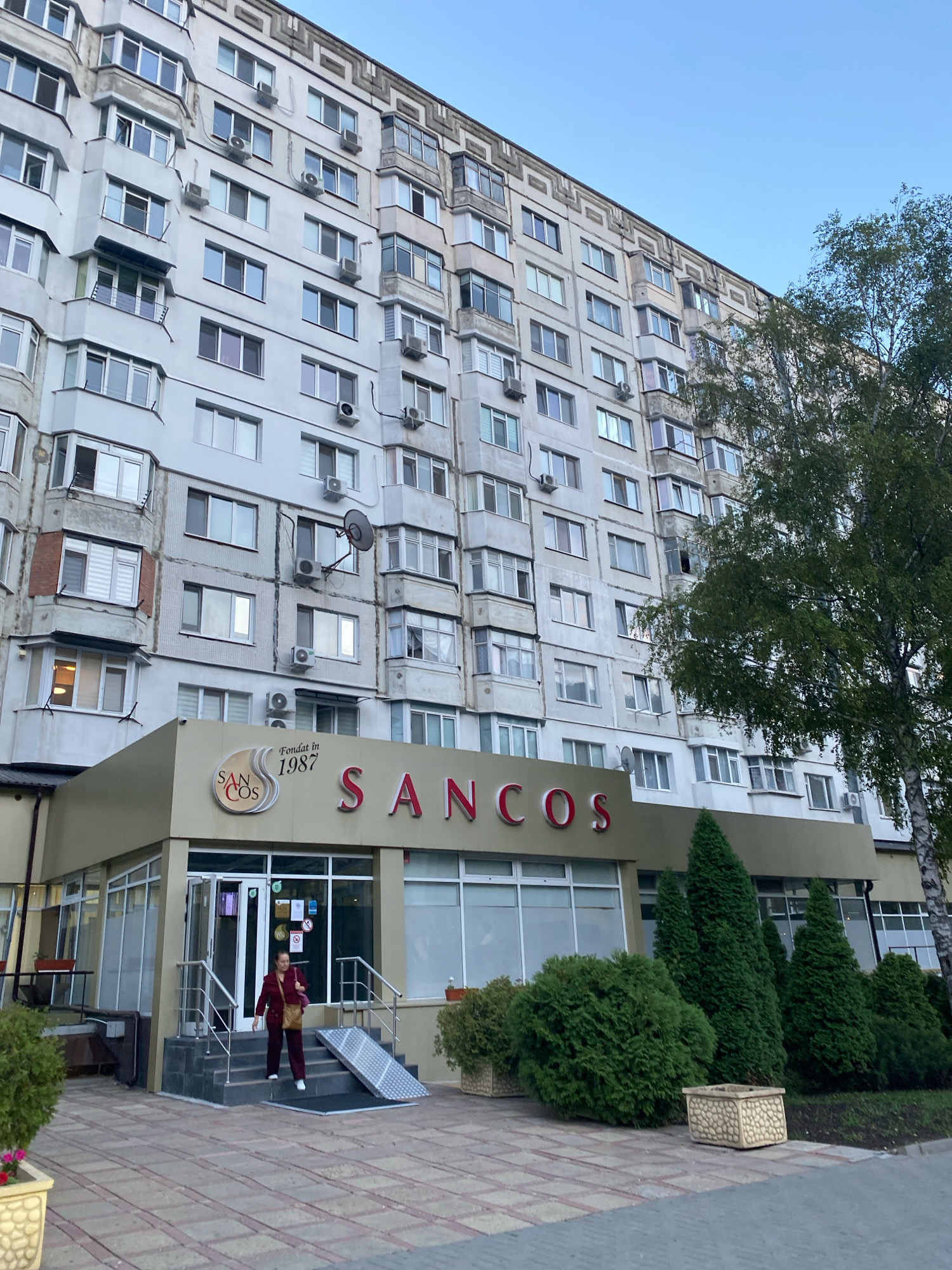The image shows a multi-story apartment building in Chișinău, Moldova. The building is grey and has numerous windows and balconies. A part of the building at ground level has a commercial facade with a sign that reads "SANCOS". A woman in a maroon outfit is walking down the steps of the building. There are trees and bushes around the building, and the sky is clear and blue. The image was taken from a slightly low angle, looking up at the building.