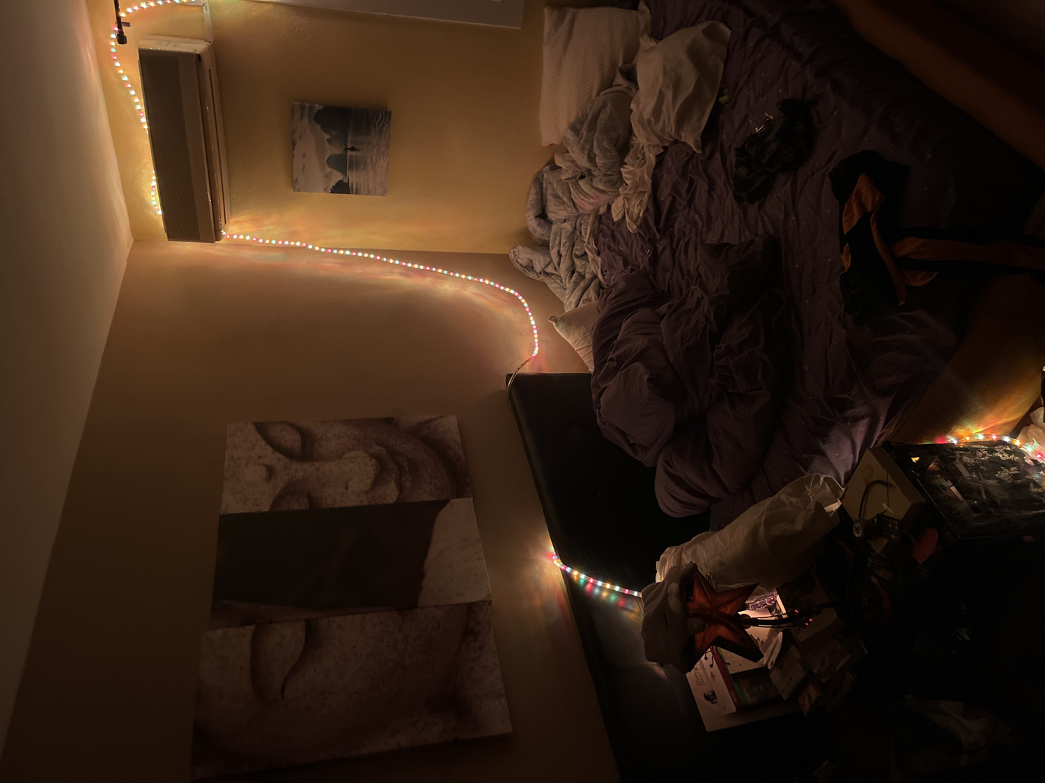 The image captures an indoor bedroom scene, likely at night or in low-light conditions, illuminated primarily by colorful LED string lights. The perspective is tilted, looking down into the room from an elevated angle.
The central focus is an unmade bed with dark purple or grey bedding, several white pillows, and what appears to be discarded clothing on top, including a dark garment and an orange fabric.
Colorful LED string lights are strung along the upper wall near the ceiling, passing over a wall-mounted white air conditioning unit. Another section of similar multi-colored string lights illuminates a dark surface next to the bed, which holds a pile of books, a pair of headphones, and a distinctive red, star-shaped ornament or light fixture.
Two pieces of wall art decorate the light brown walls. Above the air conditioning unit, there's a smaller, black-and-white picture, possibly a landscape or seascape depicting a solitary figure. On the lower wall, a larger, multi-panel artwork with a textured or abstract design in muted tones is visible.
The room is largely dim, with the string lights casting a warm, colorful glow and creating soft shadows. No people are present, and no specific activities are taking place beyond the arrangement of personal items. There is no visible text in the image. The unmade bed suggests a relaxed, lived-in space.