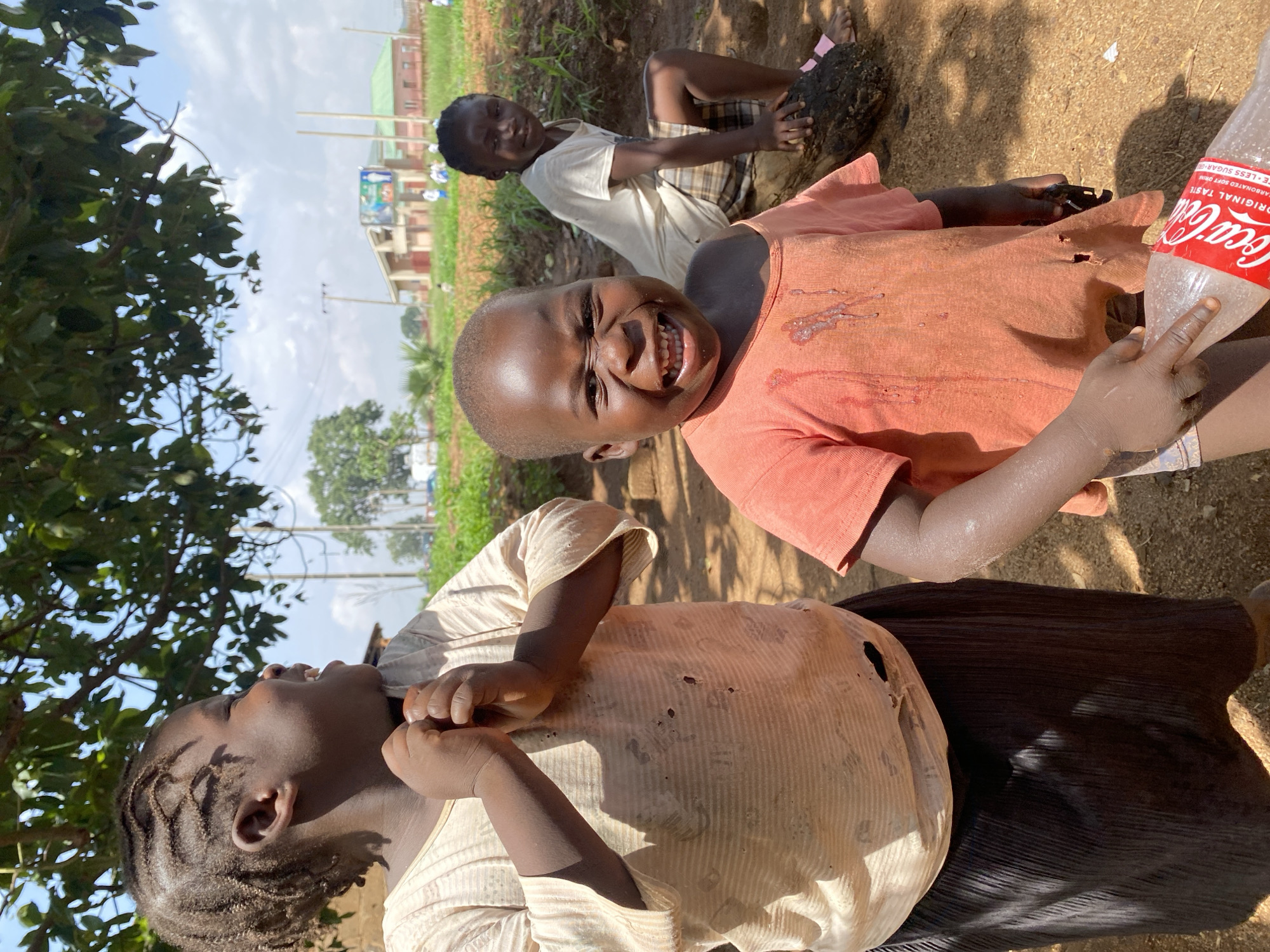 This outdoor portrait, taken in Riyom, Nigeria, features three young children under bright daylight. The sky shows scattered clouds, and the ground is dry and dusty.
In the left foreground, a child with dark, braided hair wears a light-colored, possibly cream or white, striped shirt with visible wear and holes, and dark pleated skirt or pants. This child is seen looking upwards and to the right, with bent arms.
The central subject, a child in an orange t-shirt, smiles widely and joyfully at the camera, displaying their teeth. This child holds a clear plastic Coca-Cola bottle with a red label. Splashes or residue are visible on their shirt. Text clearly visible on the bottle includes "Coca-Cola" and "ORIGINAL TASTE," with fragments of "ZE - LESS SUGAR - O" and "CARBONATED SOFT DRINK."
In the mid-ground, behind the smiling child, another child is seated or lying down, wearing a light-colored shirt and plaid shorts. This child looks towards the right, near a dark, possibly rubber or tire-like object.
The background features green foliage and trees on the left, with additional trees, buildings (including one with a green roof), and utility poles suggesting a semi-urban or rural setting. The children appear to be engaged in play or rest outdoors. The strong sunlight casts shadows, indicating a clear day.