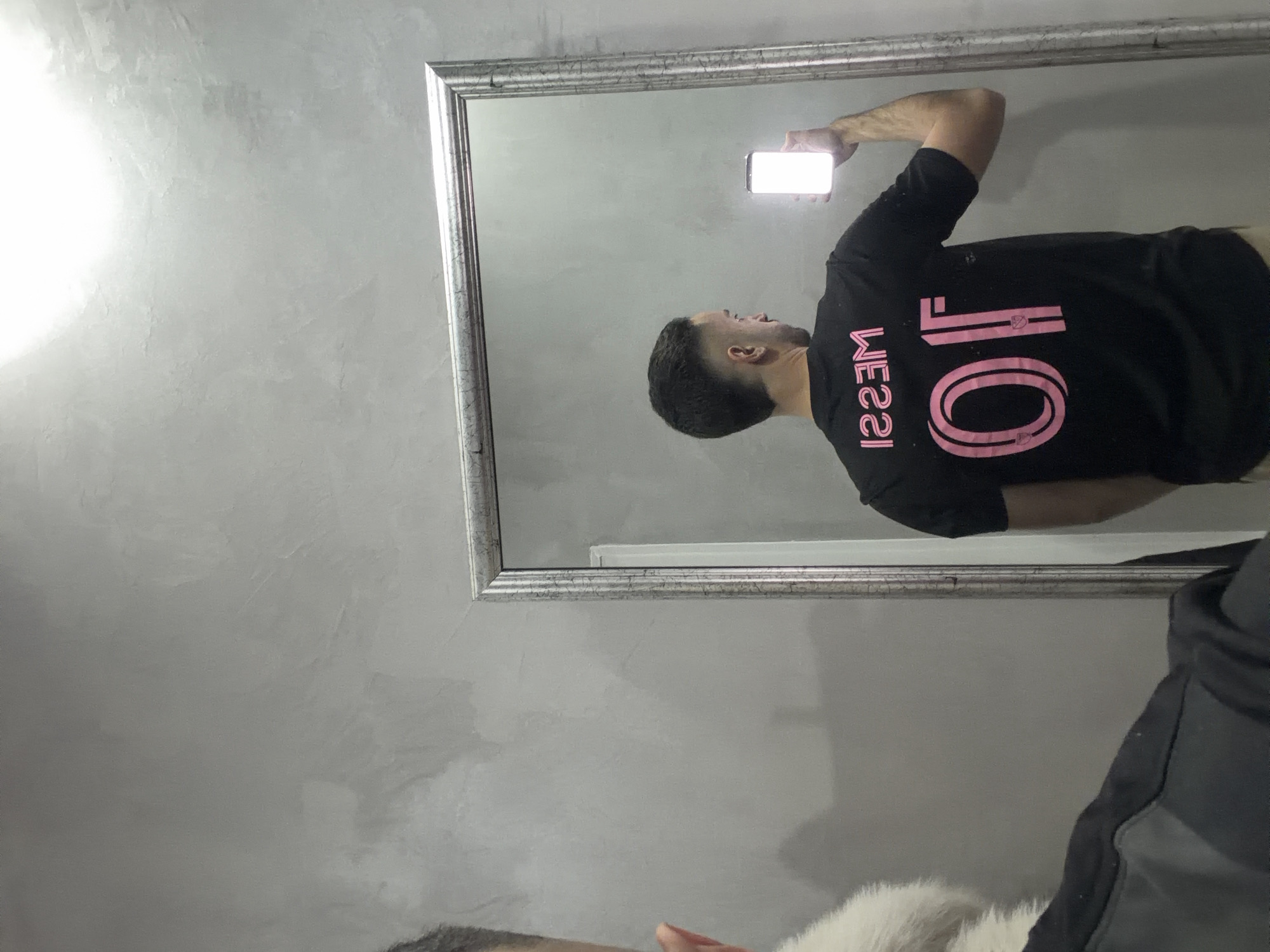A man wearing a black t-shirt with "MESSI" and the number "10" in pink is visible in a mirror. He is holding a brightly lit phone. The mirror has a silver frame and is hanging on a gray wall. Part of another person wearing a gray hoodie is seen in the lower portion of the image. The location is not evident from the image.
