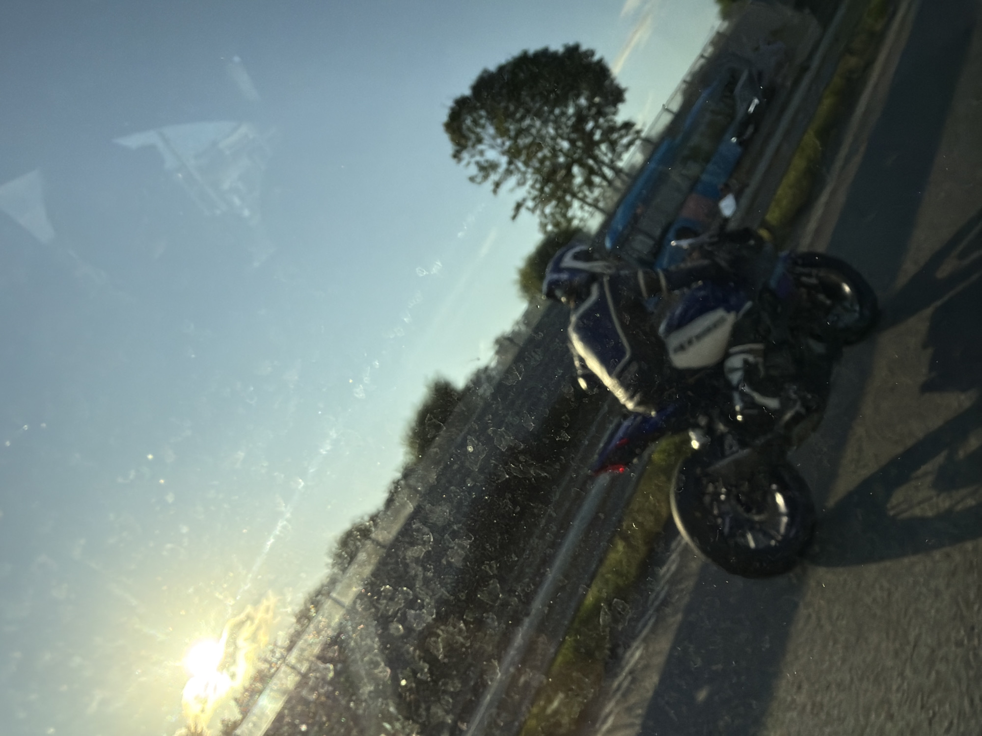 The image shows a motorcycle rider on a blue and white motorcycle, traveling on a road, likely a highway, with a bright sun in the upper left corner. The rider is wearing a helmet and protective gear. In the background, there are trees and a blue bus. The scene appears to be captured from inside a vehicle, as evidenced by the reflections and dirt on the glass. The image is tilted, giving a dynamic feel to the motion. The location is identified as Gothenburg, Sweden, suggesting a setting within or near the city.