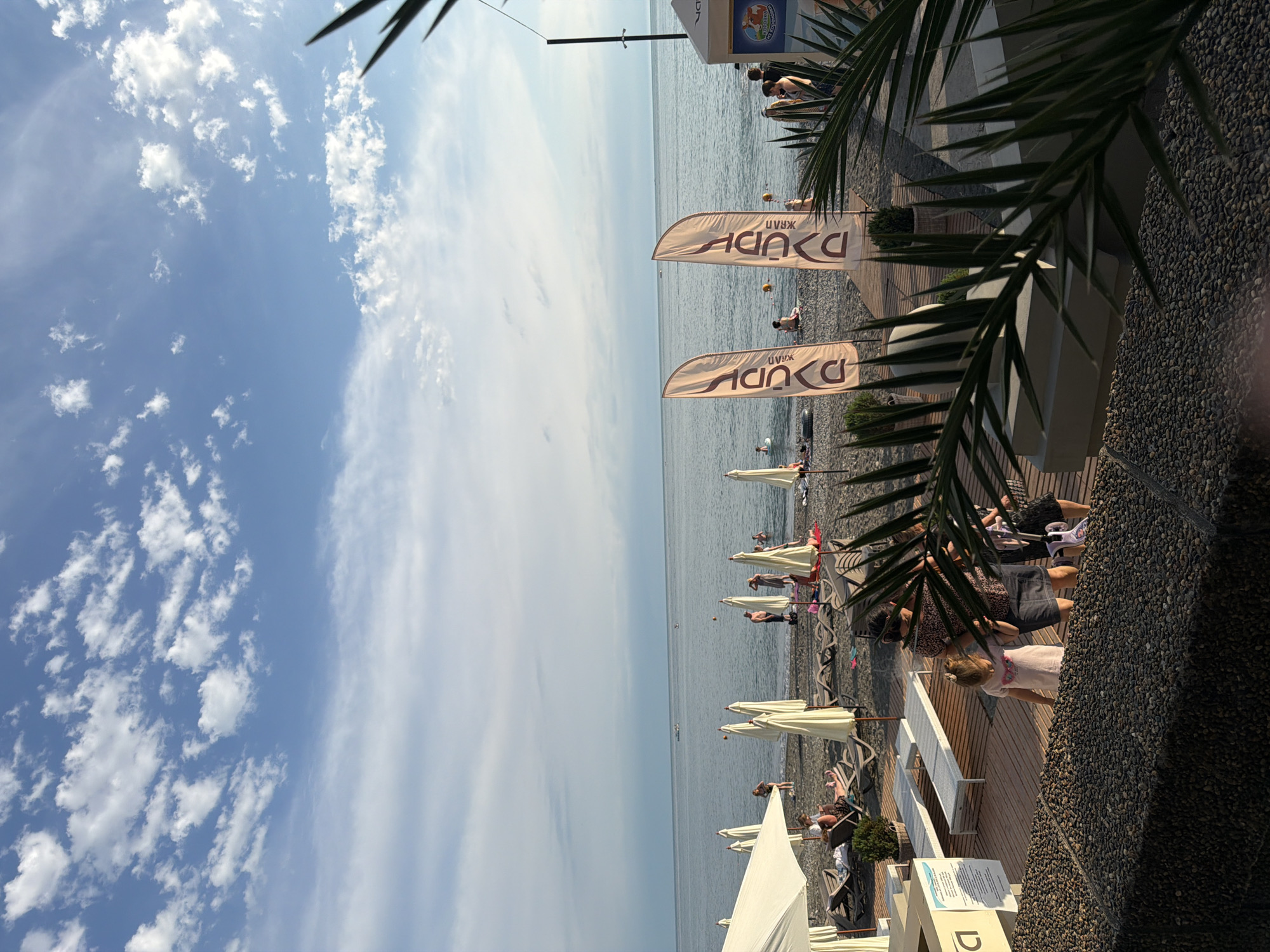 The image depicts a sunny day at a beach resort in Sochi, Russia. The scene is populated with beachgoers relaxing under umbrellas and near the water. The beach itself is made of dark pebbles, characteristic of the Sochi coastline. In the foreground, palm fronds partially obscure the view, suggesting a tropical or subtropical resort setting. Two tall, light-colored flags with text are prominently displayed, likely advertising a beach club or service. In the background, the calm sea stretches to the horizon, meeting a sky dotted with white clouds. The overall atmosphere is one of leisure and vacation.
