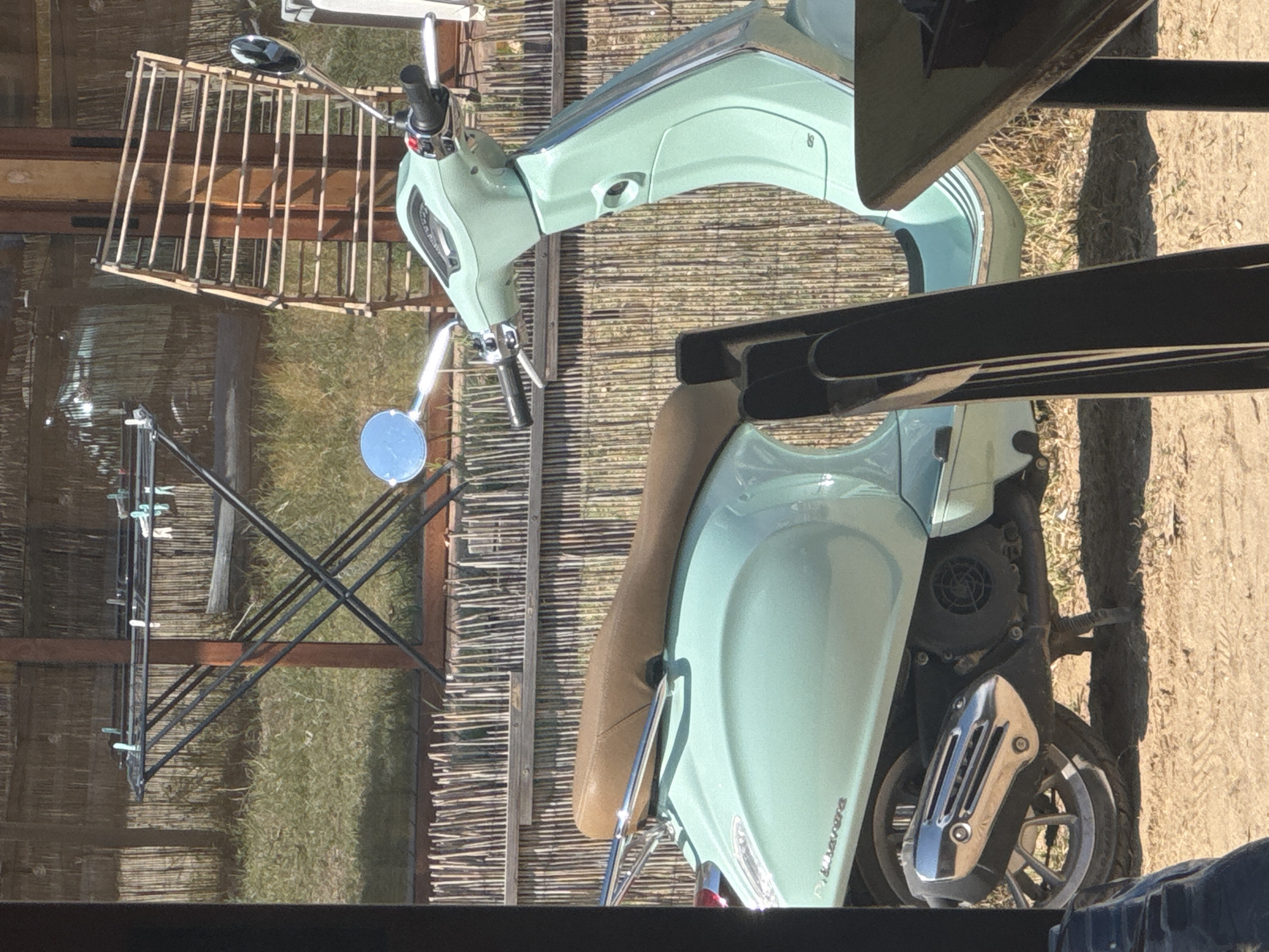 A light blue Vespa scooter is parked on a dirt surface. The scooter has a brown seat and chrome accents. Behind the scooter, there is a bamboo fence and some drying racks. The scene appears to be outdoors. The image was taken at an unspecified location in Sozopol, Bulgaria.