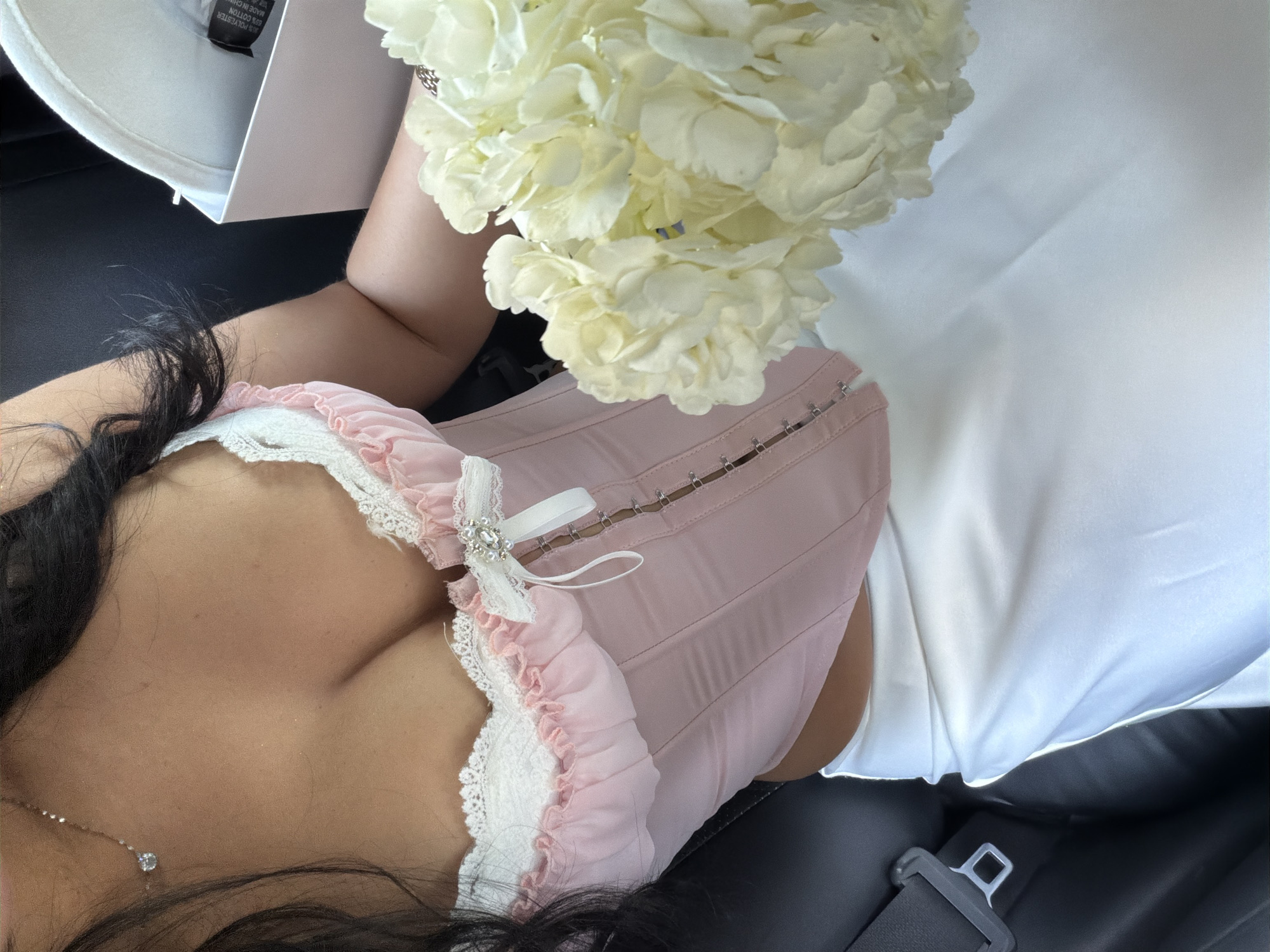 A person is lying down in what appears to be a vehicle. The person is wearing a pink and white corset-style garment. The person has dark hair, and a necklace is visible. A bouquet of white flowers is held near the person's upper body. A white box is visible in the top left corner, and a white cloth covers part of the scene. A seatbelt buckle is visible in the lower right corner. The location is not specified, and cannot be determined from the visual data.