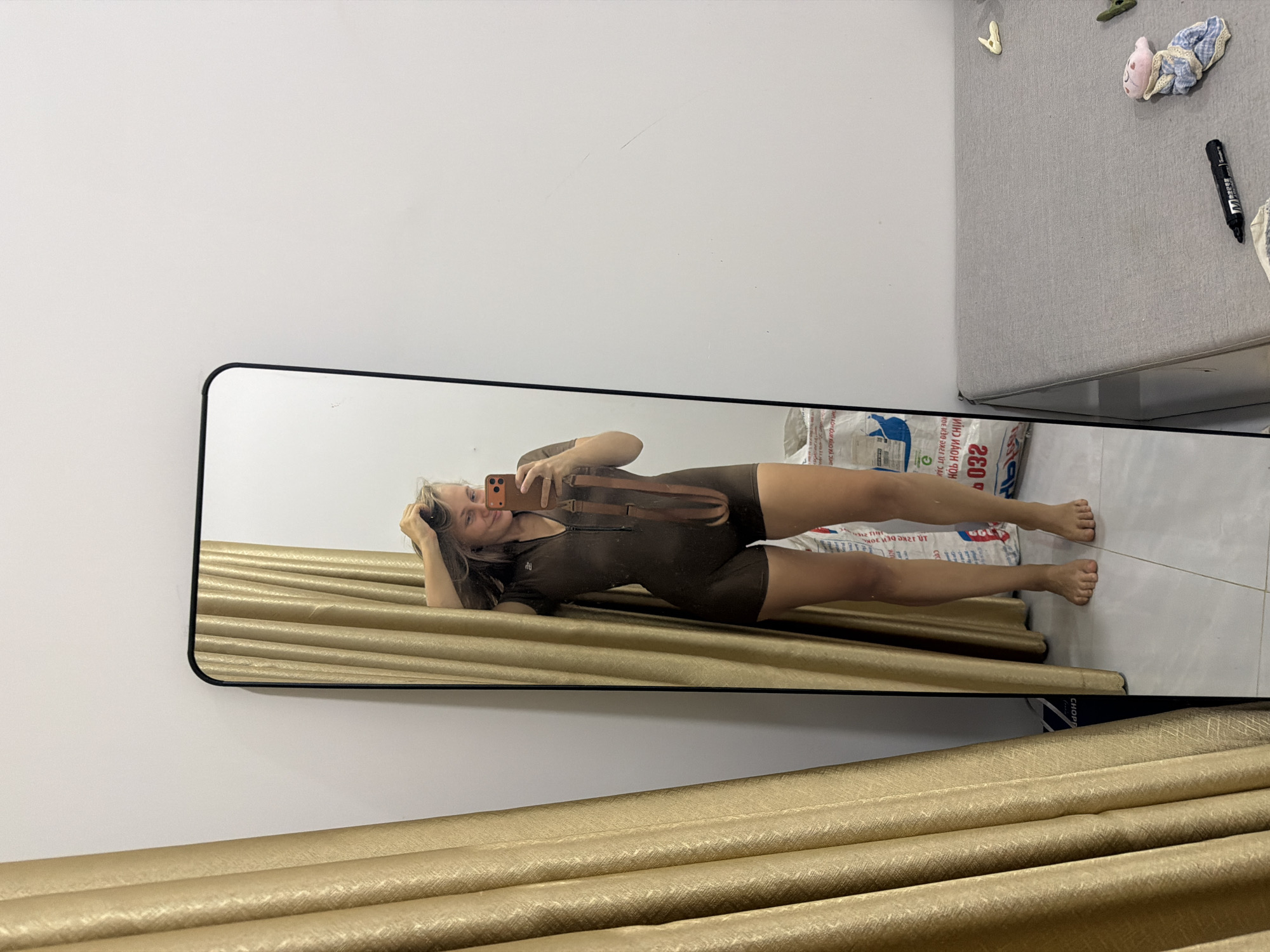 In Bac Nha Trang Ward, Vietnam, a woman stands in front of a full-length mirror, taking a selfie. She is wearing a dark brown, short-sleeved, high-neck jumpsuit with a zipper down the front. Her right hand is raised, touching her hair, and she holds an orange smartphone in her left hand. A brown strap, possibly from a bag, is draped across her chest and shoulder.
The mirror reflects her from the waist up, showing her blonde hair and smiling face. Behind her, to the left, is a golden curtain with a textured pattern. To the right of the mirror, the wall is white and appears to have some faint scratches. In the background, behind the woman's reflection and to the right of the mirror, are two large white bags with text and red numbering visible.
The floor is tiled with a light-colored pattern. A portion of a gray upholstered sofa is visible in the lower right corner of the frame, with a black marker and a small fabric doll lying on it.