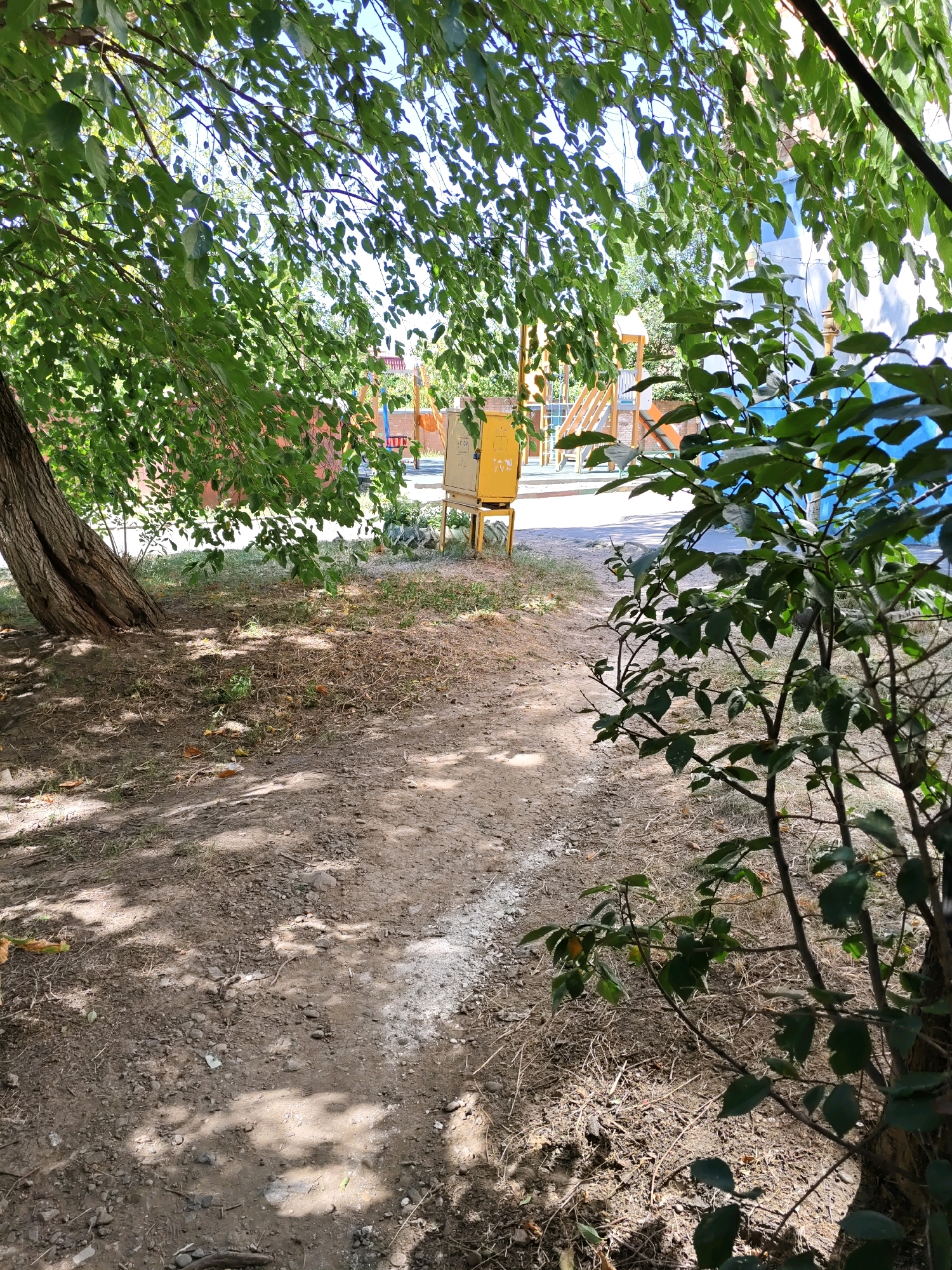 The image captures an outdoor scene on a sunny day, likely in a residential courtyard or park area within the city of Rostov-on-Don, Russia. In the foreground, a rough dirt path with scattered dry grass and pebbles is visible. Sunlight filters through the dense green foliage of a large tree on the left, casting dappled shadows on the ground.
Further into the scene, a yellow metal utility box or cabinet stands prominently. Behind it, a playground structure with colorful slides and platforms can be seen, suggesting a family-friendly environment. The background includes a building with a light blue and white facade. To the right, a young tree with green leaves partially obscures the view of the playground and building. The overall impression is one of a casually maintained urban outdoor space. No people are present in the image.