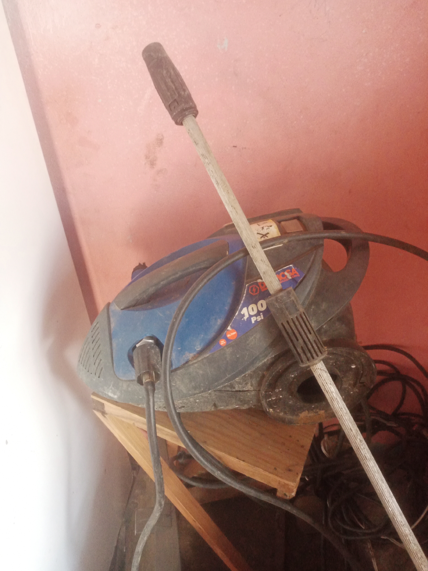 A blue and black pressure washer is seen resting on a wooden shelf against a pink wall in La Tagua, Venezuela. The pressure washer has a silver wand with a black handle extending upwards and to the left, partially obscuring the machine. Black power cords and hoses are visible behind and around the pressure washer, suggesting it is connected or ready for use. The overall scene is indoors, with a white wall visible on the left and the pink wall extending to the right. The lighting suggests an indoor environment with some ambient light.