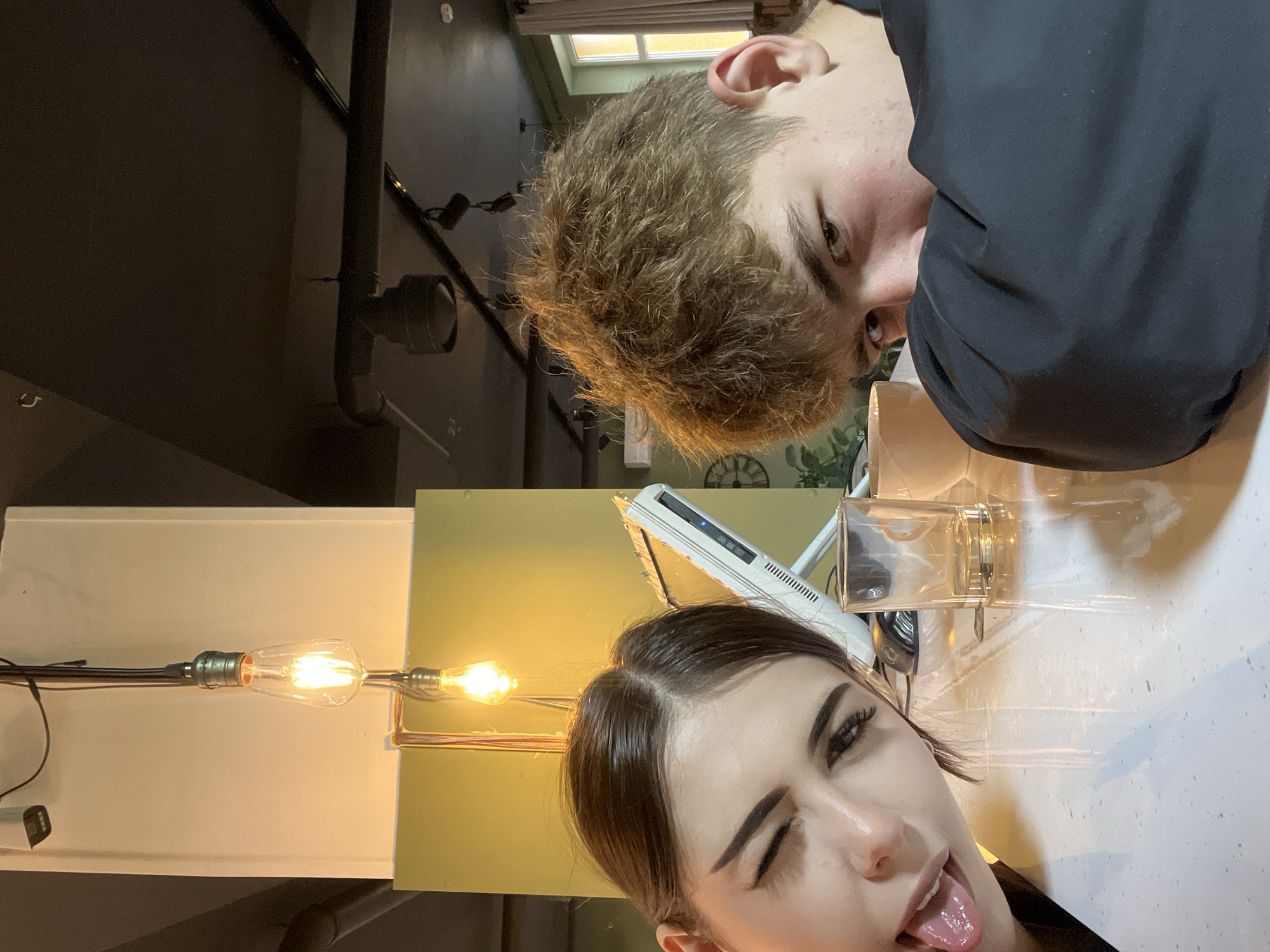 An indoor scene in a cafe located in Красные Баки, Russia, captures two young individuals in a close-up, slightly Dutch-angled shot. On the left, a female with dark hair winks with her right eye and protrudes her tongue towards the camera. On the right, a male with light brown, disheveled hair looks towards the camera, partially obscuring his face with the dark sleeve of a long-sleeved garment. On the white speckled table between them are an empty clear glass tumbler and a light-colored coffee cup. In the mid-ground, a white all-in-one computer monitor with a visible disc drive is present. The background features light beige and yellowish-green walls, with a dark ceiling from which exposed pipes and track lighting fixtures are suspended. Two prominent Edison-style bare light bulbs hang from wires, emitting warm light. A white security camera is mounted high on the left wall. A window with white frames and a partially visible large, dark-faced clock are seen on the right side of the frame, along with some green plant foliage below the window.