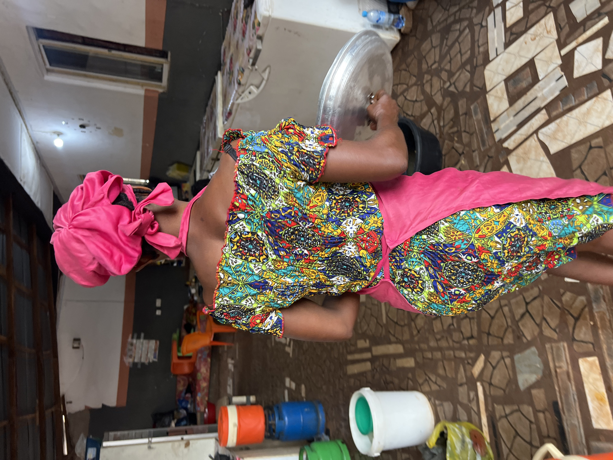 A woman, viewed from the back, is wearing a pink headwrap and a vibrantly patterned, off-the-shoulder top with puff sleeves. A pink apron is tied around her waist, extending down to her knees. She is holding a large, round metal lid in her right hand. The woman is standing on a tiled floor with an irregular, mosaic-like pattern. In the background, there are several large white refrigerators, a blue barrel topped with an orange container, and other miscellaneous items, suggesting a market or a food preparation area. The scene appears to be indoors, possibly in Nkpor, Nigeria, given the context.