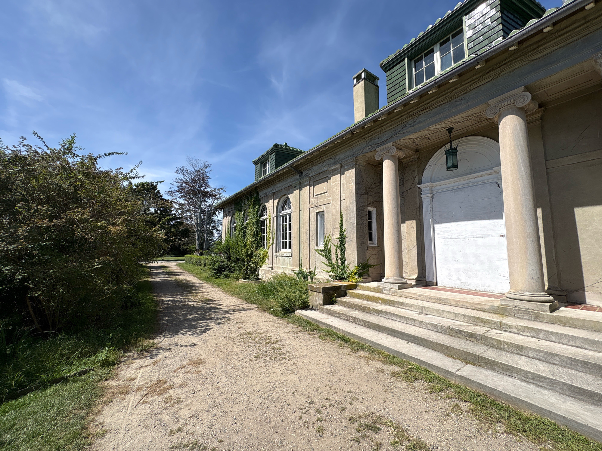 The image captures a side view of a large, old stone building with a green, shingled roof and dormer windows. The building features classical architectural elements such as large columns, arched windows, and a prominent entrance with a lantern. A gravel path leads away from the building, bordered by lush green bushes and trees. The sky is a clear blue with a few wispy clouds. The scene suggests a well-maintained historical estate or public building, possibly within a park or garden setting, located in New London, United States of America. The lighting indicates it is daytime, with sunlight casting shadows. No people are visible in the frame.