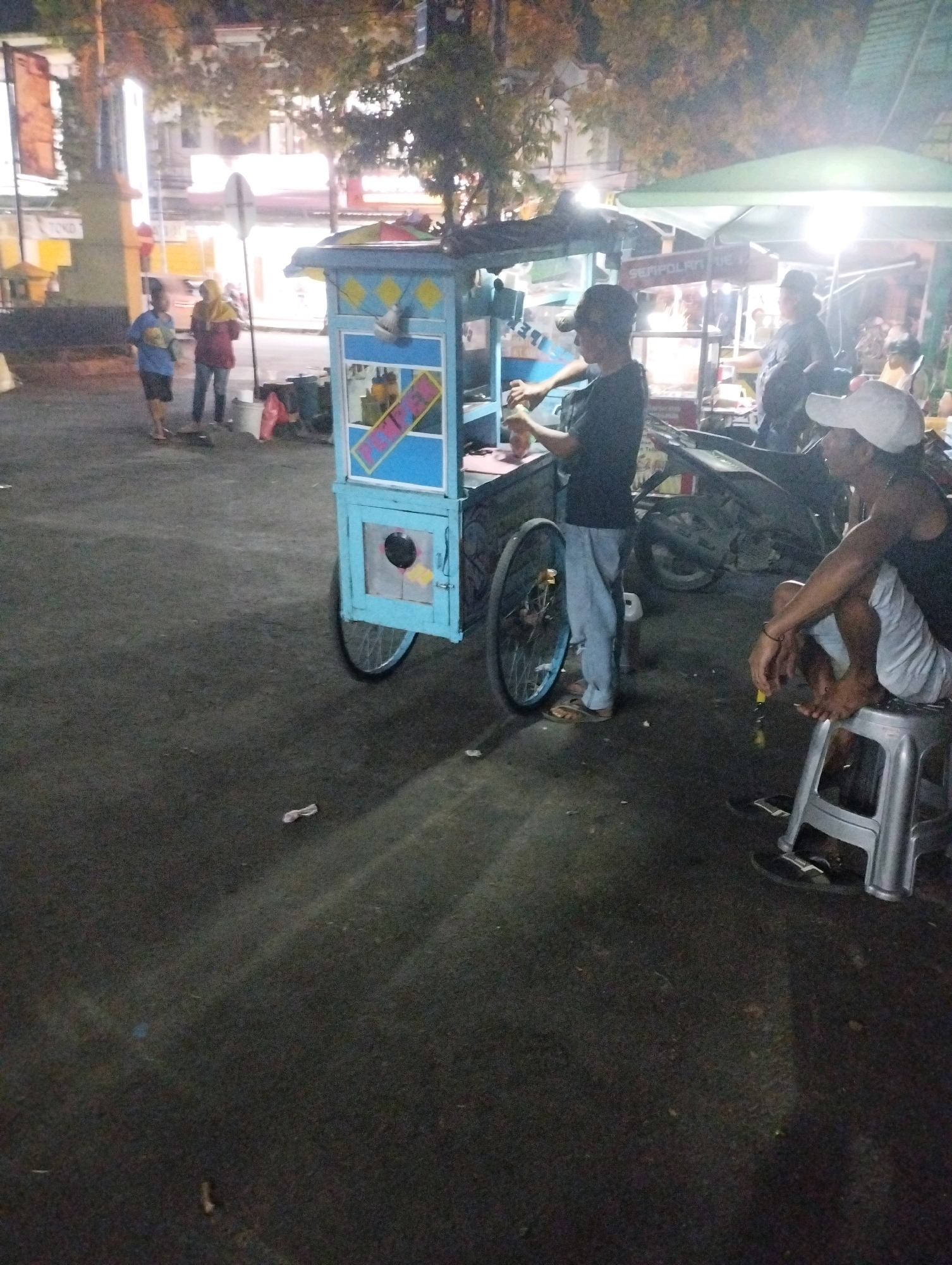 In Kecamatan Pati, Indonesia, an outdoor night scene depicts a food cart with a blue and yellow painted exterior, adorned with diamond patterns and text. A young person, wearing a blue shirt and dark shorts, stands behind the cart, engaged in a transaction. To the right, another person, dressed in a black tank top and white cap, sits on a silver stool, facing away from the cart. Behind them, a motorcycle is parked. In the background, two individuals, one in a yellow hijab and maroon top, and another in a blue shirt, stand near a tall pole with a white sign. The ground is paved and appears to be a street or market area, with some scattered debris. The lighting suggests artificial illumination from the food cart and surrounding structures.