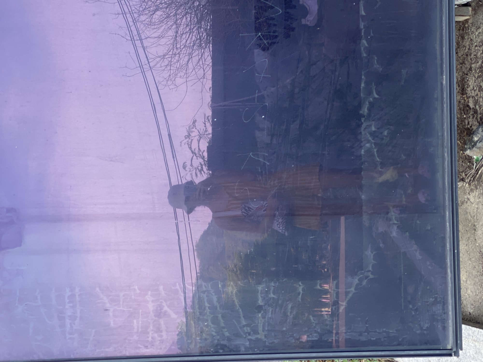 A person wearing a yellow dress and a bucket hat is reflected in a purple, dirty glass surface. The reflection shows the person holding a phone, taking a selfie, and carrying a patterned bag. The background reflected in the glass includes trees, hills, power lines, and some indecipherable markings on the glass. The scene appears to be outdoors in an urban or semi-urban setting, possibly in Bac Nha Trang Ward, Vietnam, given the presence of power lines and vegetation typical of the region. The glass is smudged and has streaks, obscuring some details of the reflection and the environment.