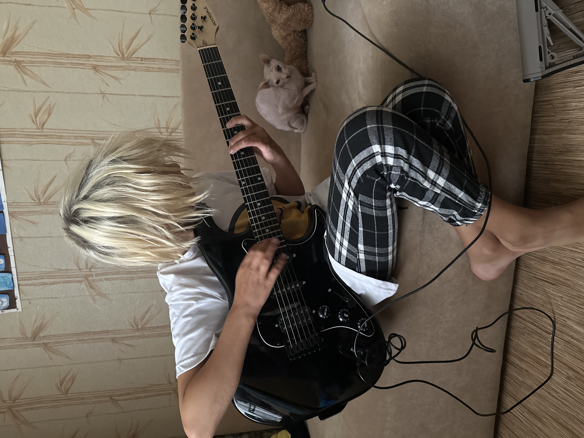 In a well-lit indoor setting, a person with blonde, disheveled hair sits on a couch, playing a black electric guitar. They are wearing a white t-shirt and black and white plaid pants. The guitar is plugged in with a black cable. To the left of the person, a section of wallpaper with a bamboo pattern is visible. To the right and slightly behind the person, a small, light-colored Sphynx cat is perched on the couch, looking curiously towards the camera. A brown teddy bear is also visible on the couch behind the cat. The lighting suggests it is daytime.