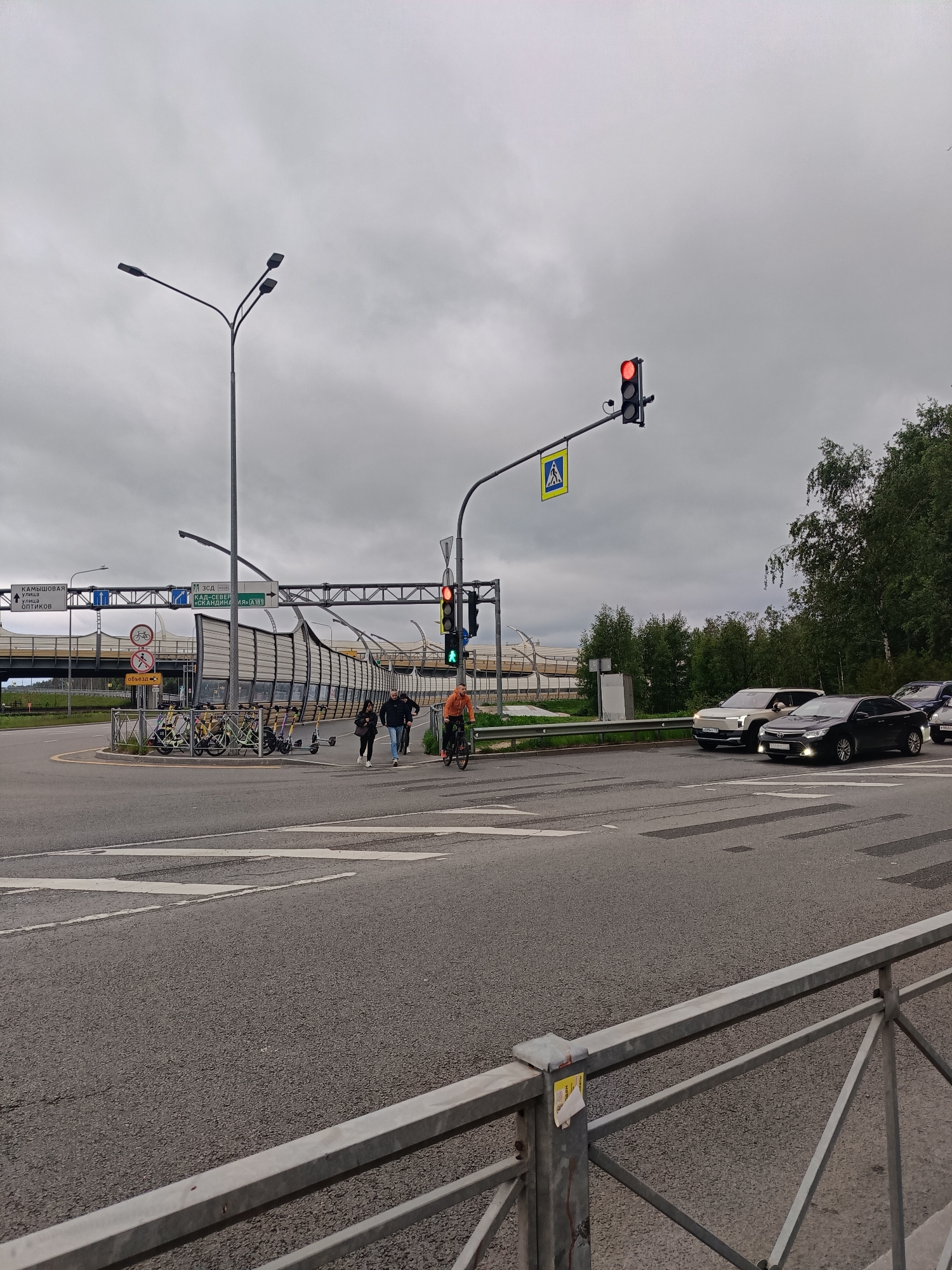The image captures a scene at a crossroads in Saint Petersburg, Russia, under an overcast sky. Several elements are present:
**Roadway and Traffic:** An asphalt roadway with crosswalk markings intersects with another road, where several cars are waiting at a red traffic light.
**Pedestrians and Cyclist:** A group of people is crossing the crosswalk, and a cyclist wearing an orange shirt is riding across the same crosswalk.
**Infrastructure:** On the side of the road are parked bicycles and scooters, as well as road signs. There are also lampposts and a sign indicating directions to the "Kadmishovaya" street, "Optics" street and "Scandinavia A181" highway.
**Environmental Features:** Trees are visible in the background. There's a metal railing in the lower part of the image. A sign with a pedestrian crossing symbol is visible.
**Overall Impression:** The image portrays a common urban traffic scene with pedestrians, cyclists, and vehicles navigating an intersection with traffic control measures.