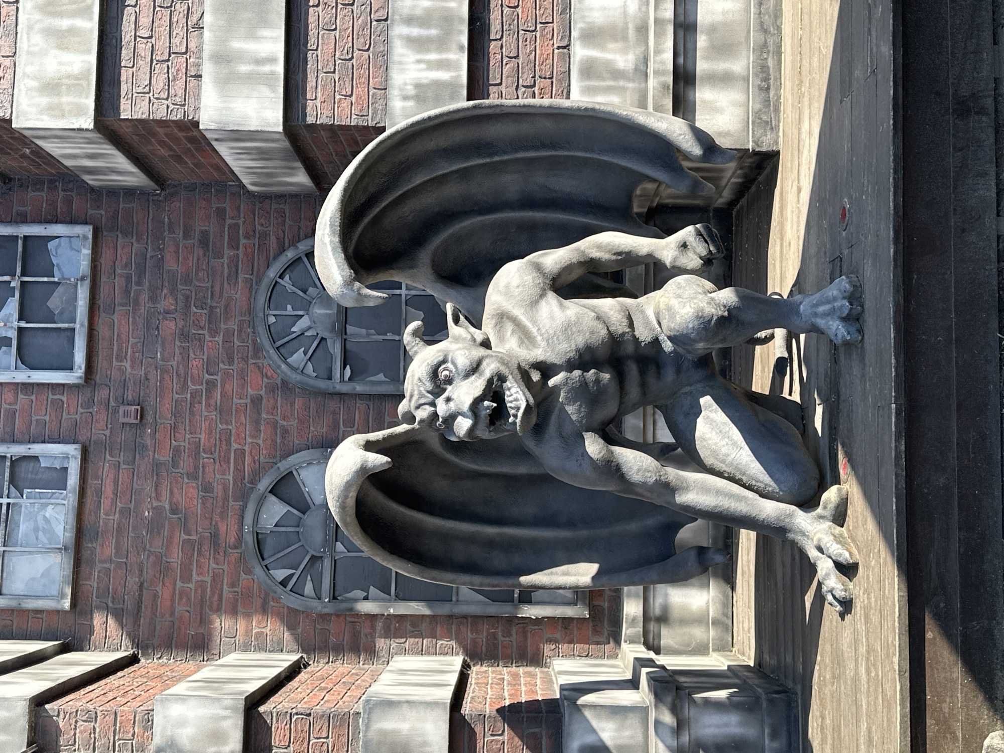 An outdoor, medium shot captures a stone statue of a winged gargoyle crouched on a wooden platform in front of a brick building. The gargoyle is positioned centrally, facing forward, with its large, leathery wings spread open behind it. Its body is muscular and reptilian, with sharp claws on its hands and feet, a pointed tail, and a monstrous head with a gaping maw filled with sharp teeth, beady eyes, and horns. The building behind the gargoyle has a red brick facade with two arched, broken windows, and grey stone architectural elements. The lighting suggests it is daytime, with strong shadows cast by the gargoyle and building. The overall scene appears to be part of a themed attraction or exhibit.