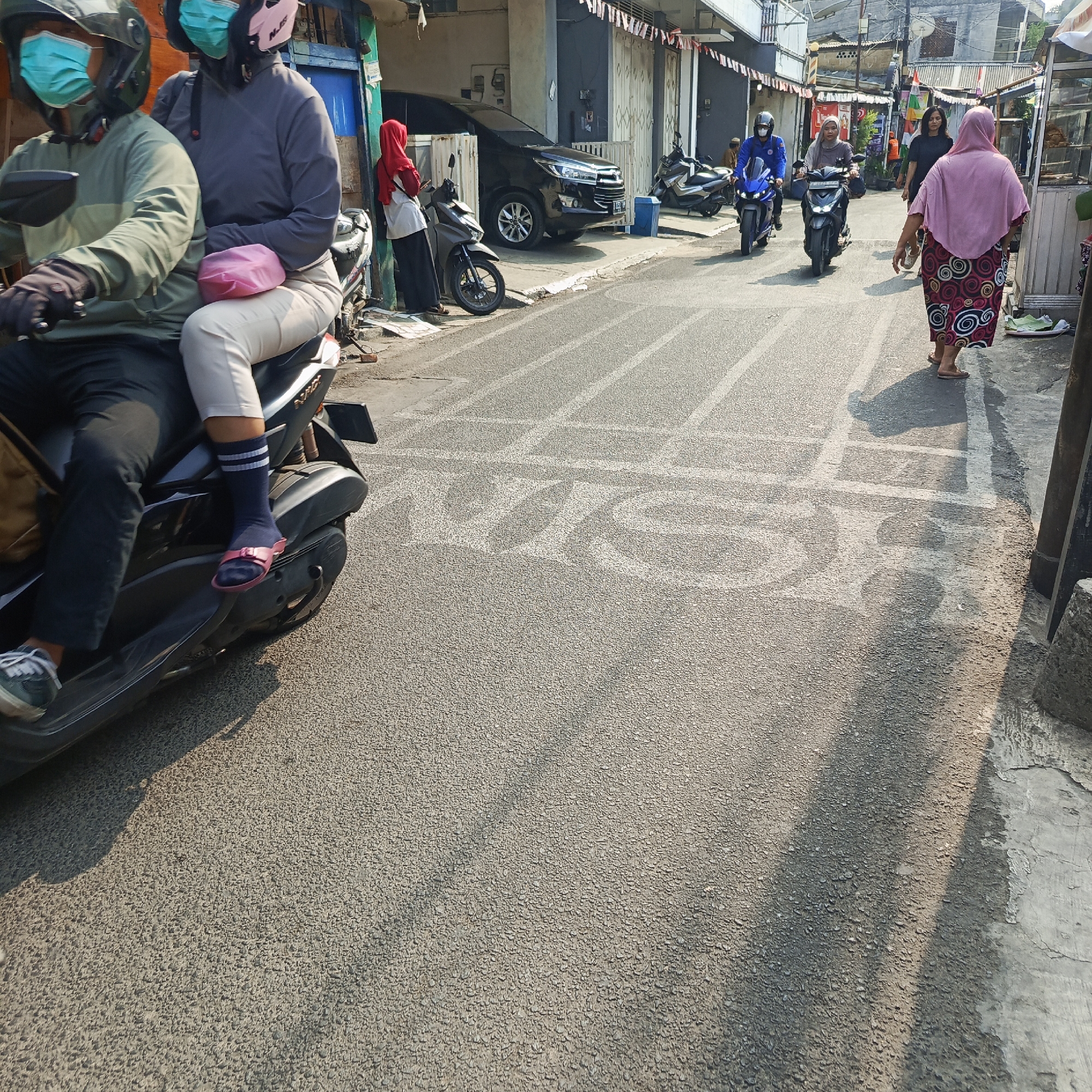 In South Jakarta, Indonesia, on a sunny day, two individuals are riding a scooter. The person in front wears a green jacket, black pants, gloves, a green helmet, and a light blue face mask. The person behind them wears a gray long-sleeved shirt, light-colored pants, a pink helmet, and a light blue face mask. A pink pouch is strapped between them.
Further down the street, another scooter carries a person wearing a gray hijab and blue face mask. A blue scooter approaches from the opposite direction with a rider in a blue jacket, helmet, and blue face mask. A woman in a pink hijab and patterned skirt walks on the right side of the road, casting a shadow. The street is paved with asphalt, and white lines are visible on the road, forming rectangular shapes. Some of these lines appear to spell out words, though they are partially obscured. Buildings line the street, some with white doors and windows, and decorative flags are visible. A black SUV is parked in front of one of the buildings.