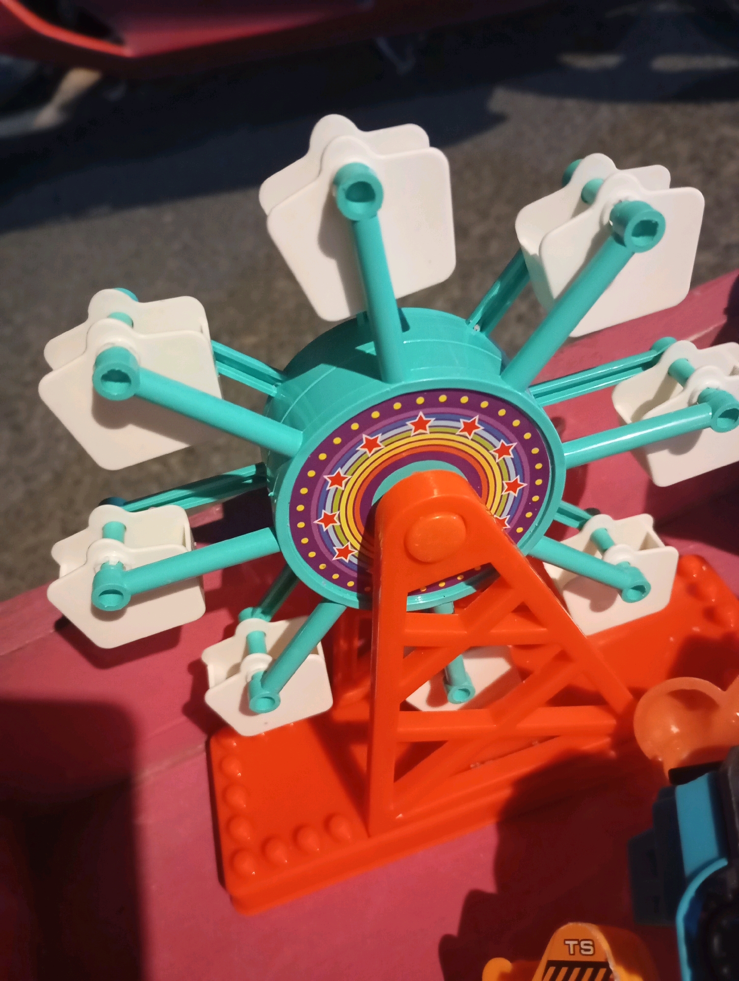 A toy Ferris wheel, primarily teal and white, stands on an orange base. The Ferris wheel has eight white gondolas attached to teal arms radiating from a central hub decorated with a rainbow and stars. The orange base has a triangular support structure. In the lower right, a blue and black toy vehicle is partially visible, with a yellow toy piece labeled "TS" in front of it. The scene appears to be outdoors on a paved surface, with a shadow cast from the Ferris wheel. The image is a close-up, high-angle shot of the toy. There is no visible indication of the location being in Randegan, Indonesia.